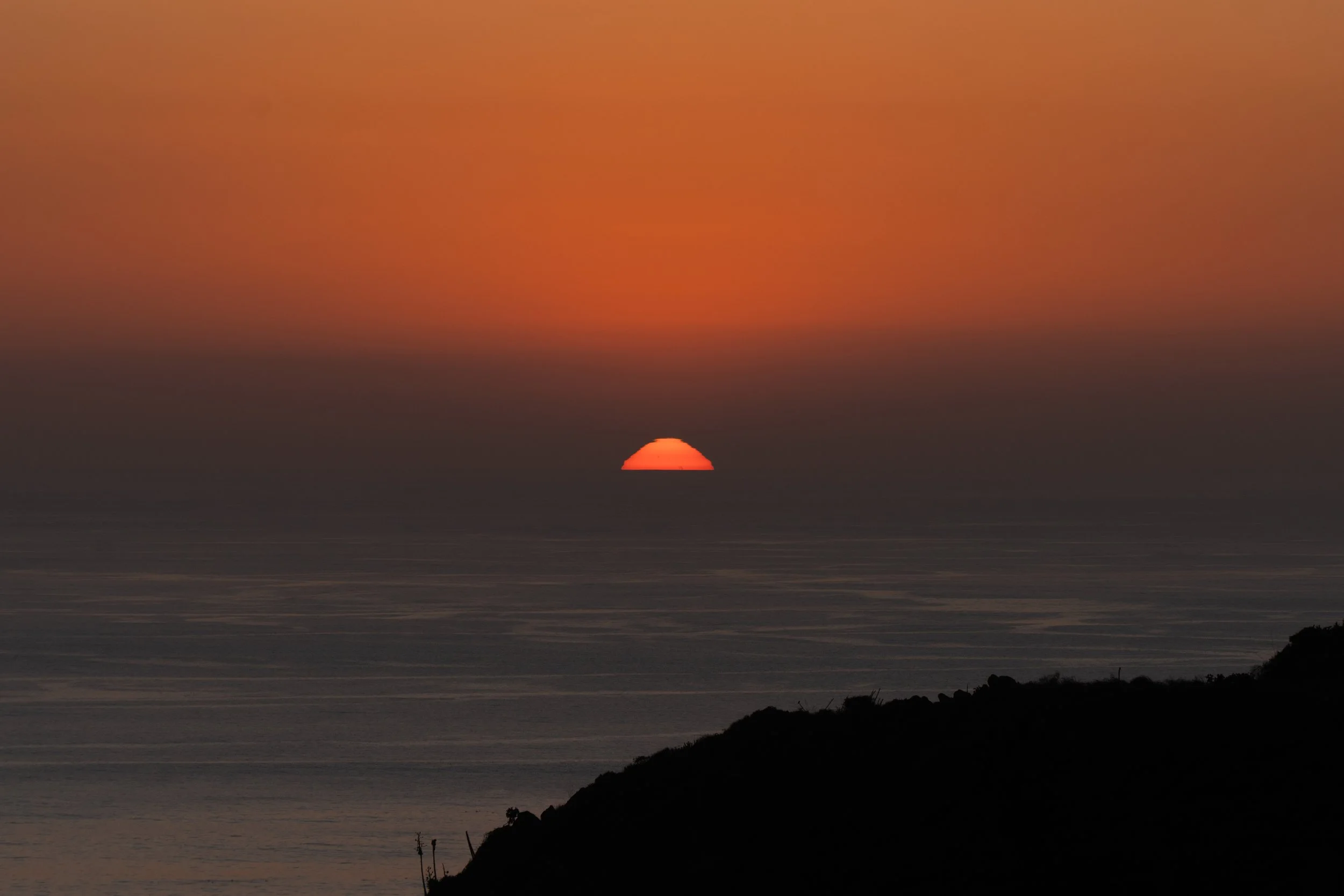 Sunset over the ocean with the sun partially below the horizon, silhouetted coastline in the foreground, and a colorful sky with shades of orange and dark clouds.