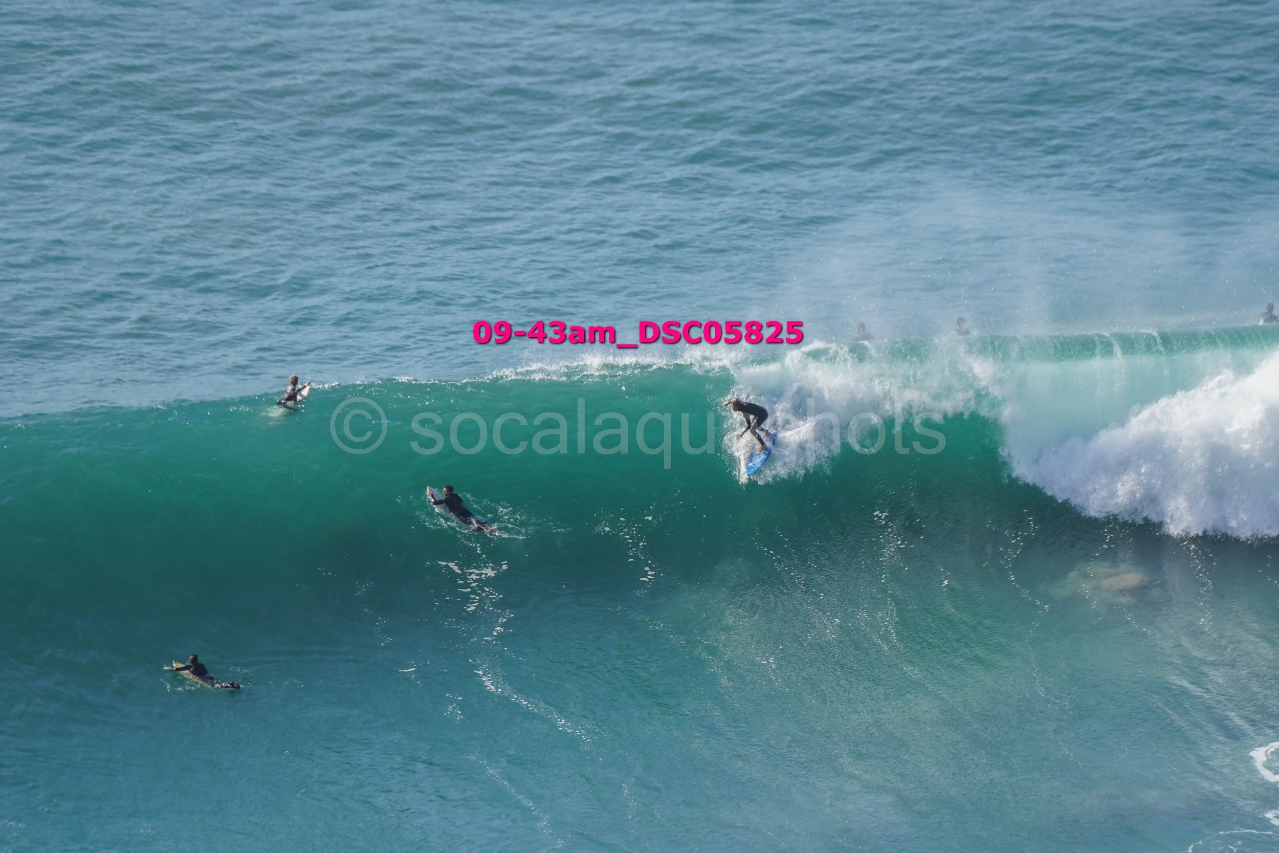 Surfer riding a large wave with several other surfers in the water nearby.