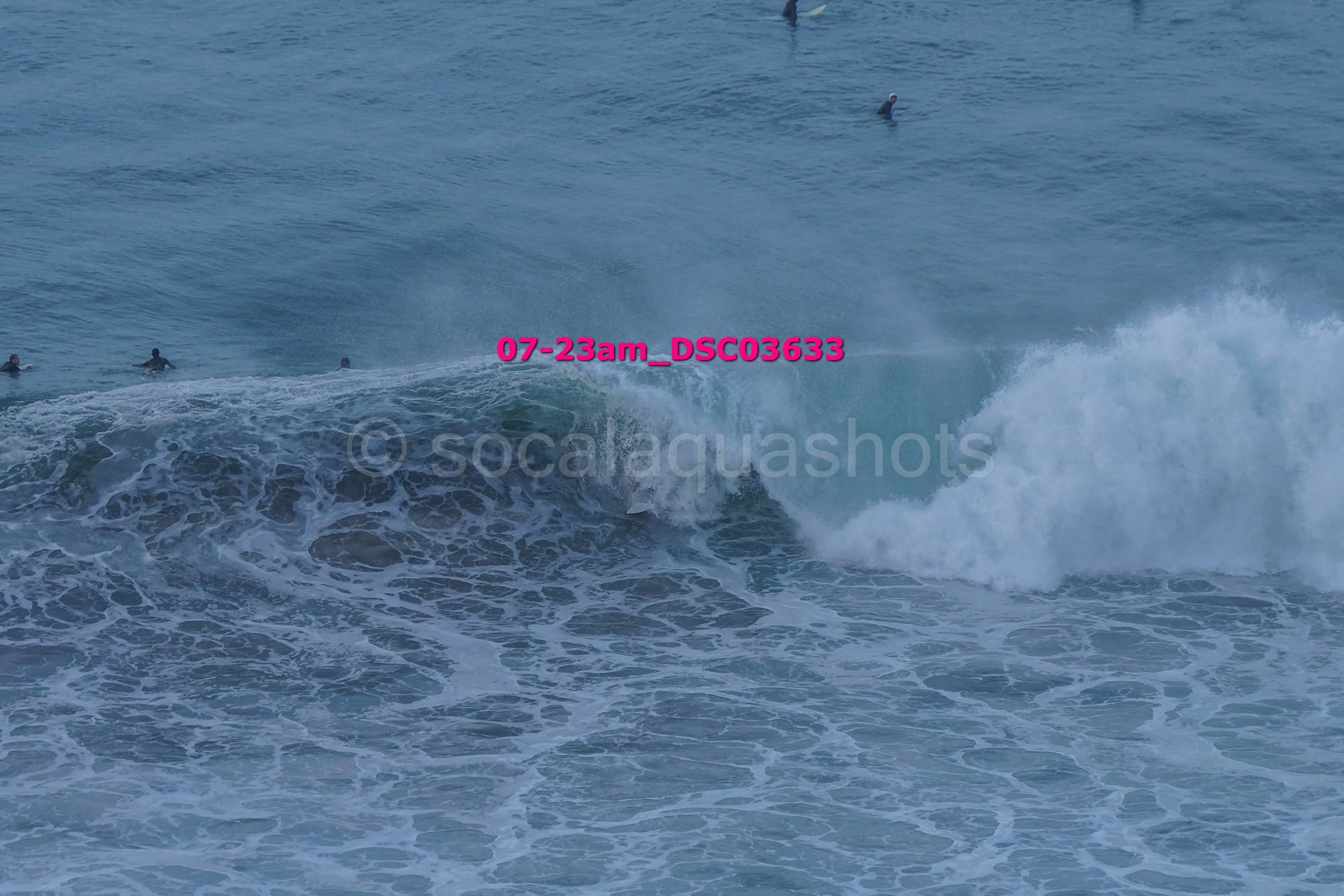 Waves crashing in the ocean with several surfers in the water.