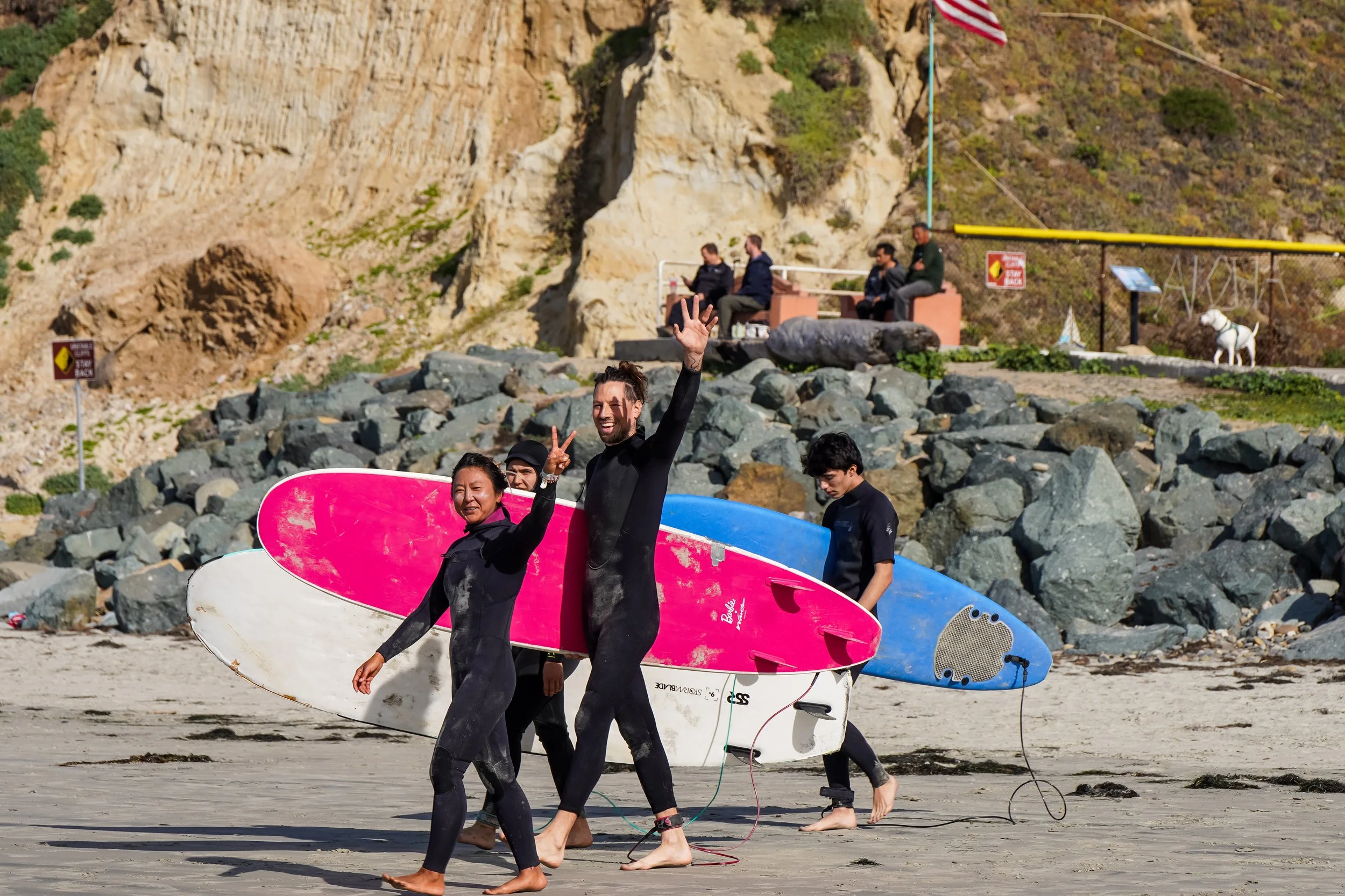 Group of people in wetsuits walking on the beach, carrying surfboards and smiling at the camera, with a rocky shoreline and cliffs in the background.