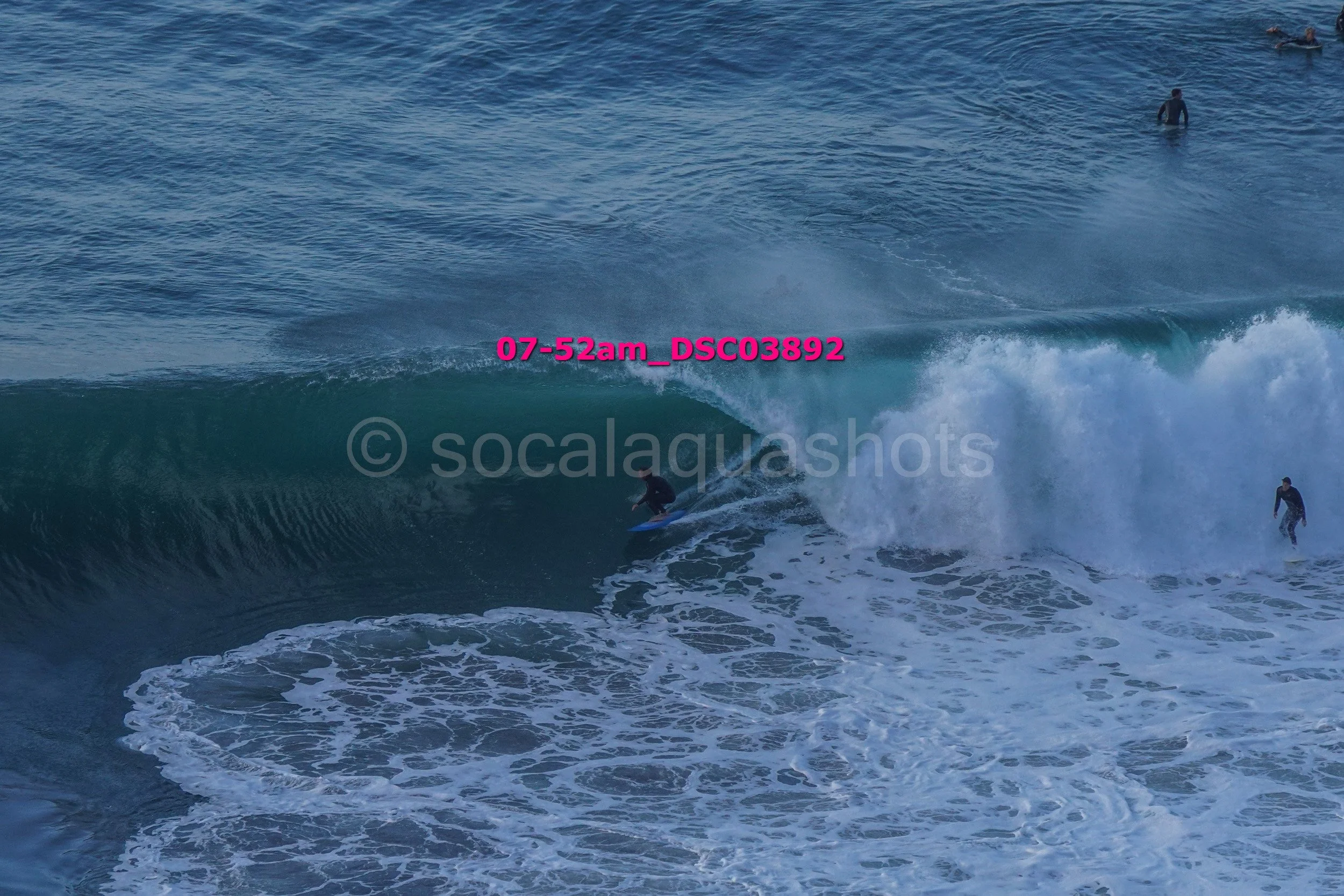 A surfer riding a large wave in the ocean with other surfers in the background.
