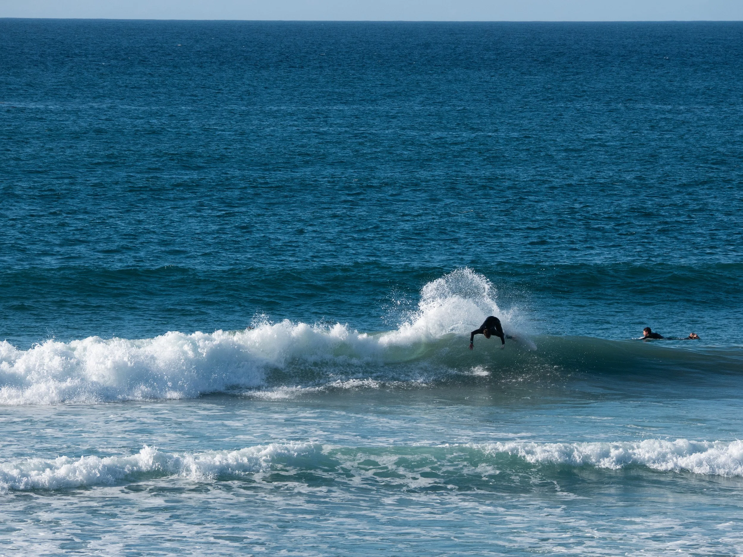Surfer performing a trick on a wave at the beach with two other surfers paddling nearby.