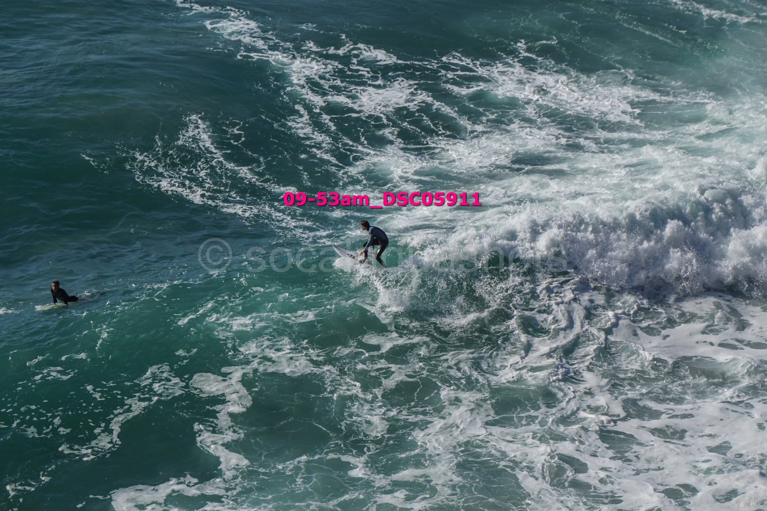 A person in a wetsuit surfing on a wave with another person lying on a surfboard nearby in the ocean.