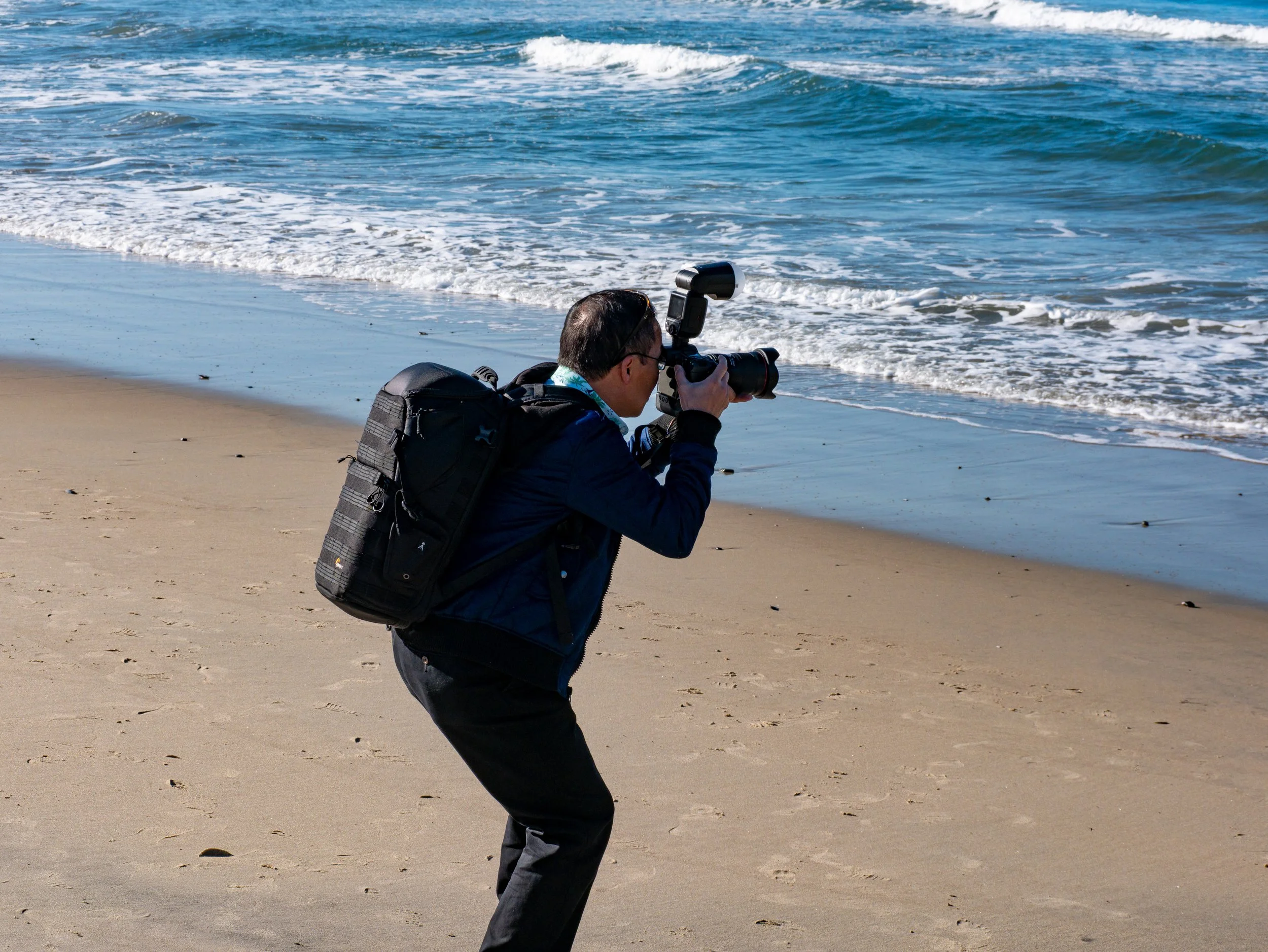 A man with a backpack taking a photograph of the ocean shoreline with a camera on a beach.