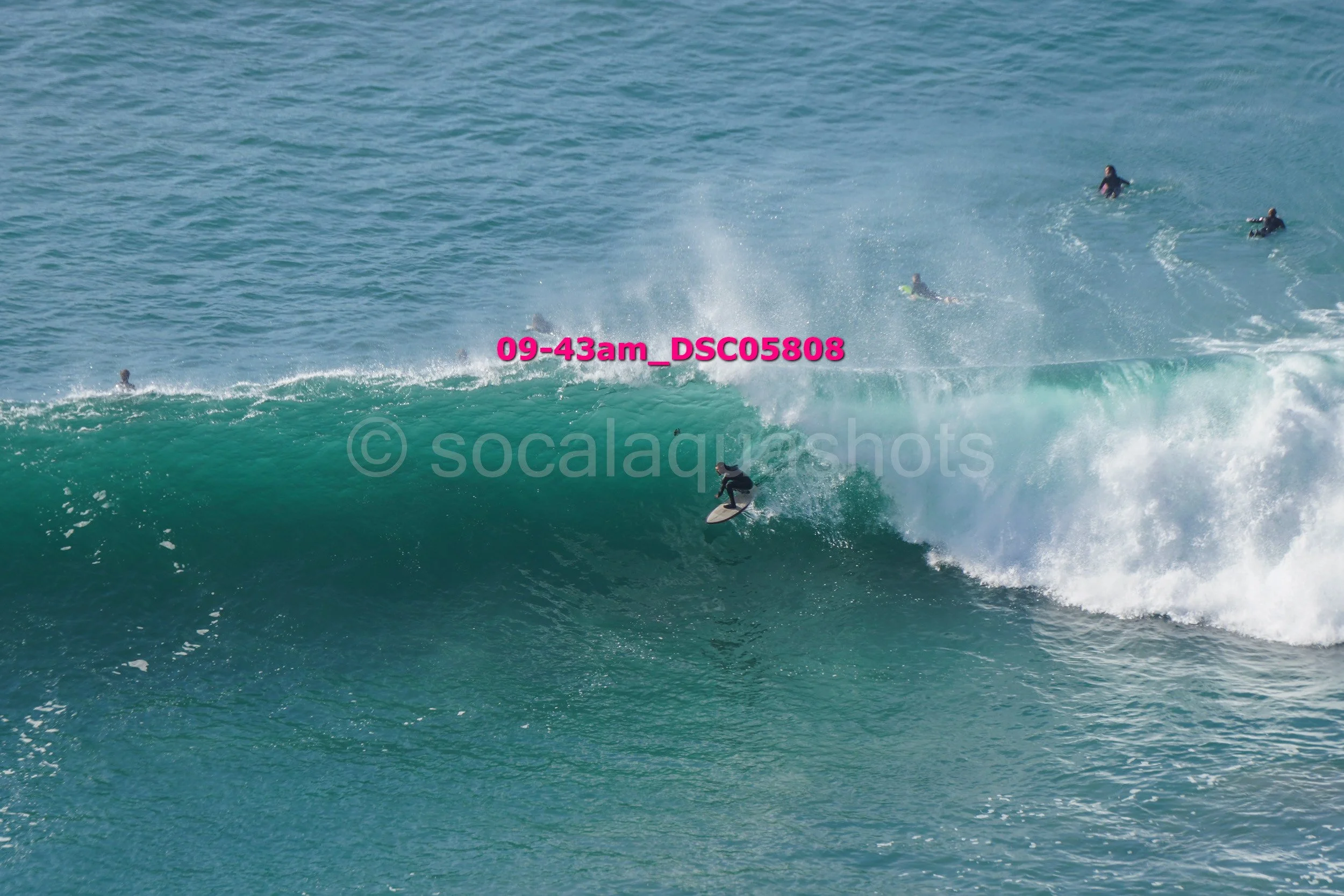 Surfer riding a large wave with several other surfers in the water nearby.
