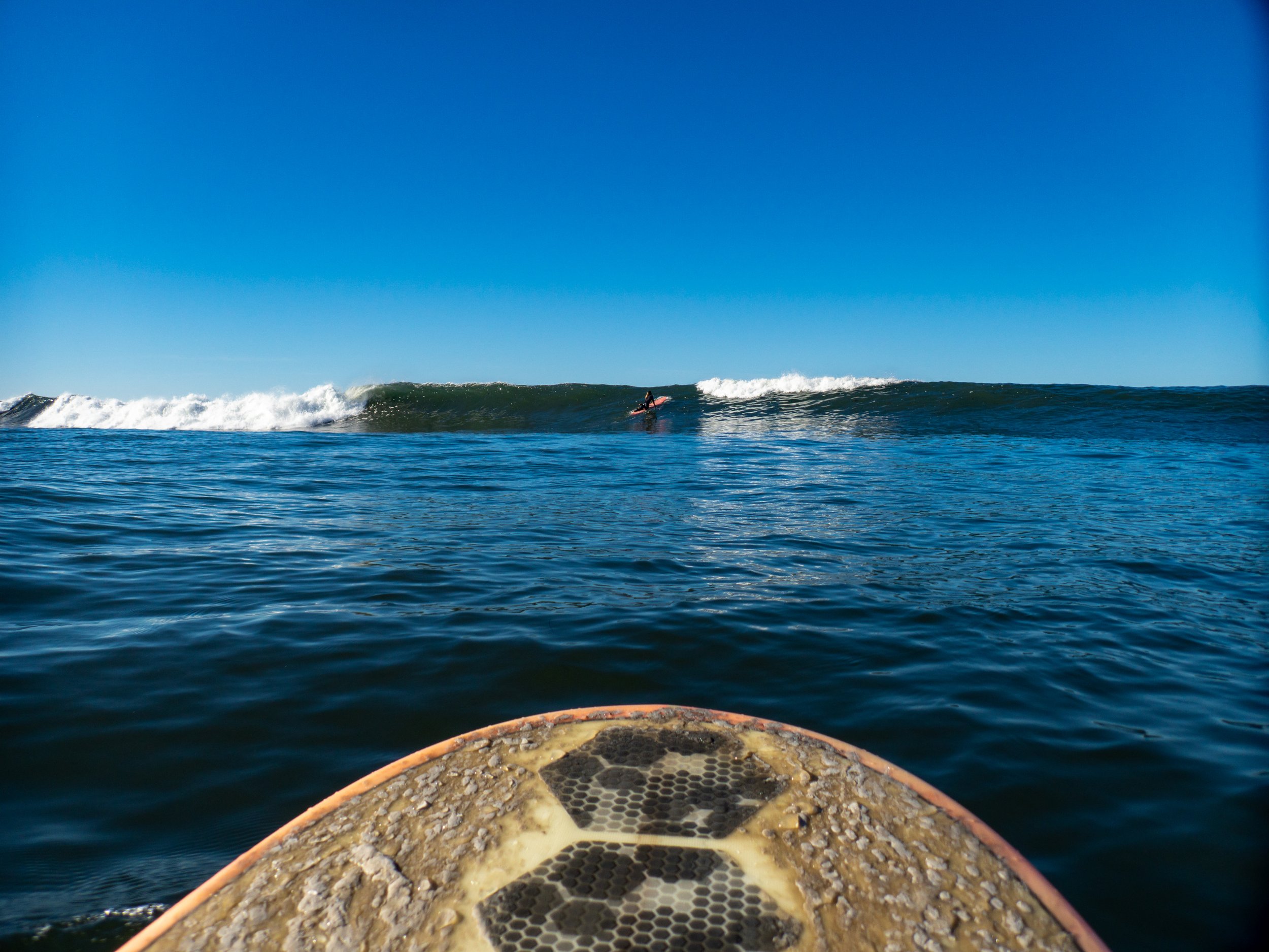 View from a surfboard looking at a surfer riding a wave in the ocean under a clear blue sky.