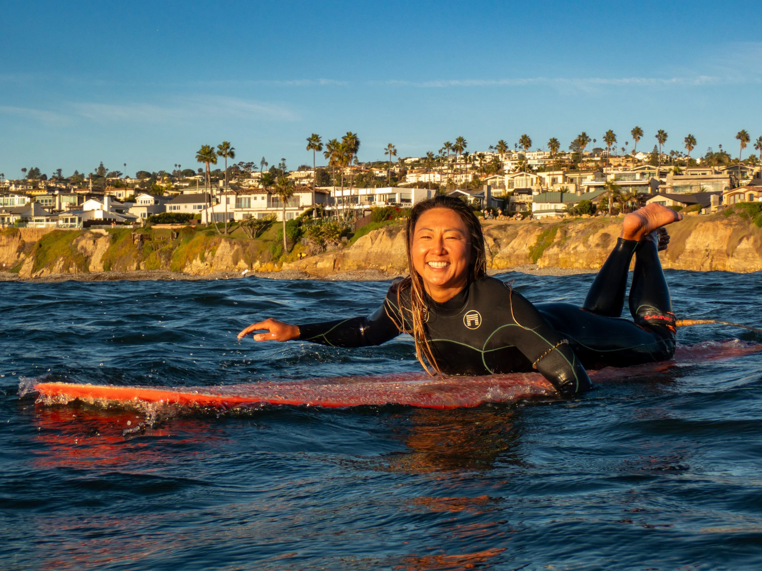 Smiling woman lying on a surfboard in the ocean near the coastline with houses and palm trees in the background.