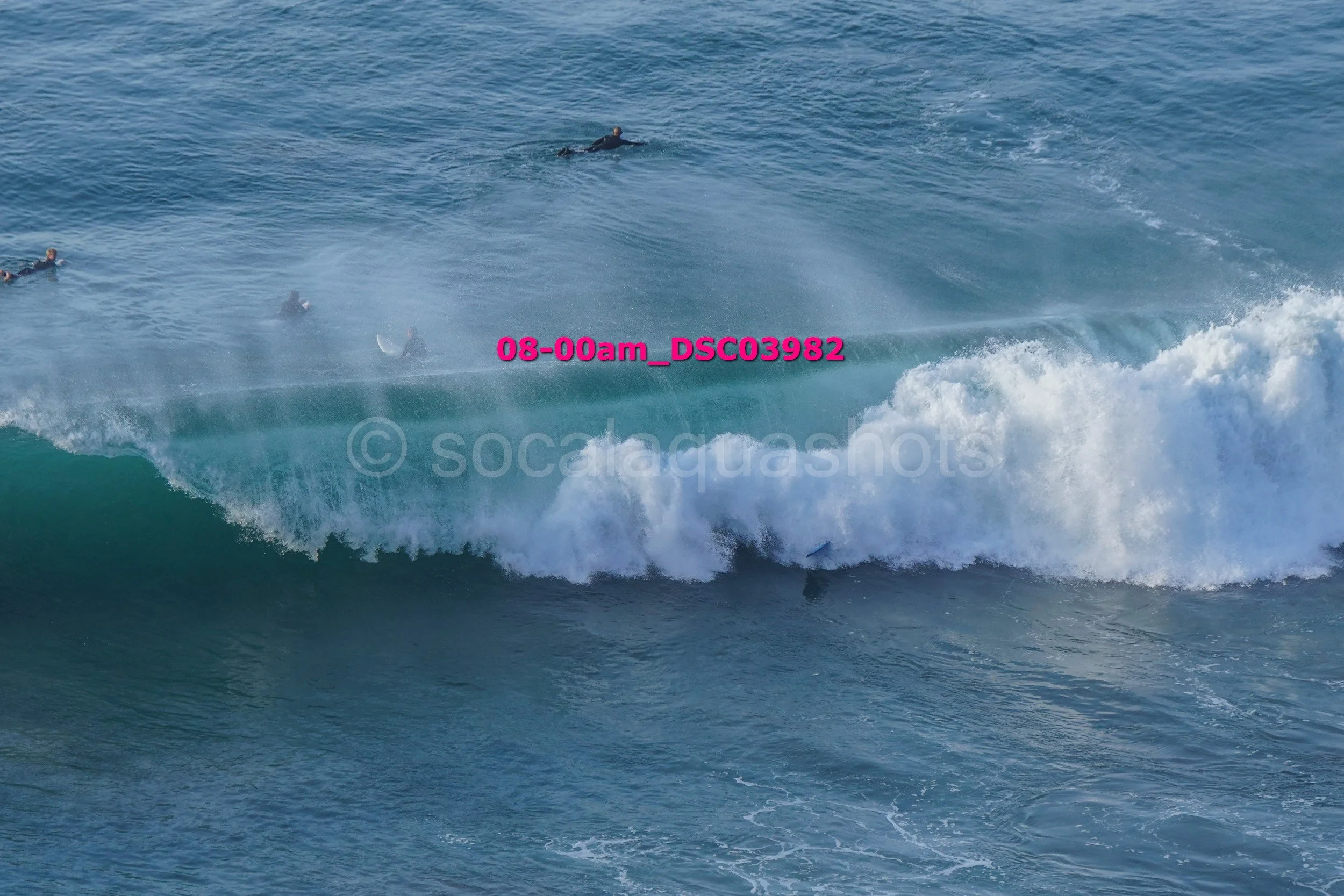 Surfers in a large ocean wave with one surfer riding inside the wave and others paddling nearby in the water.