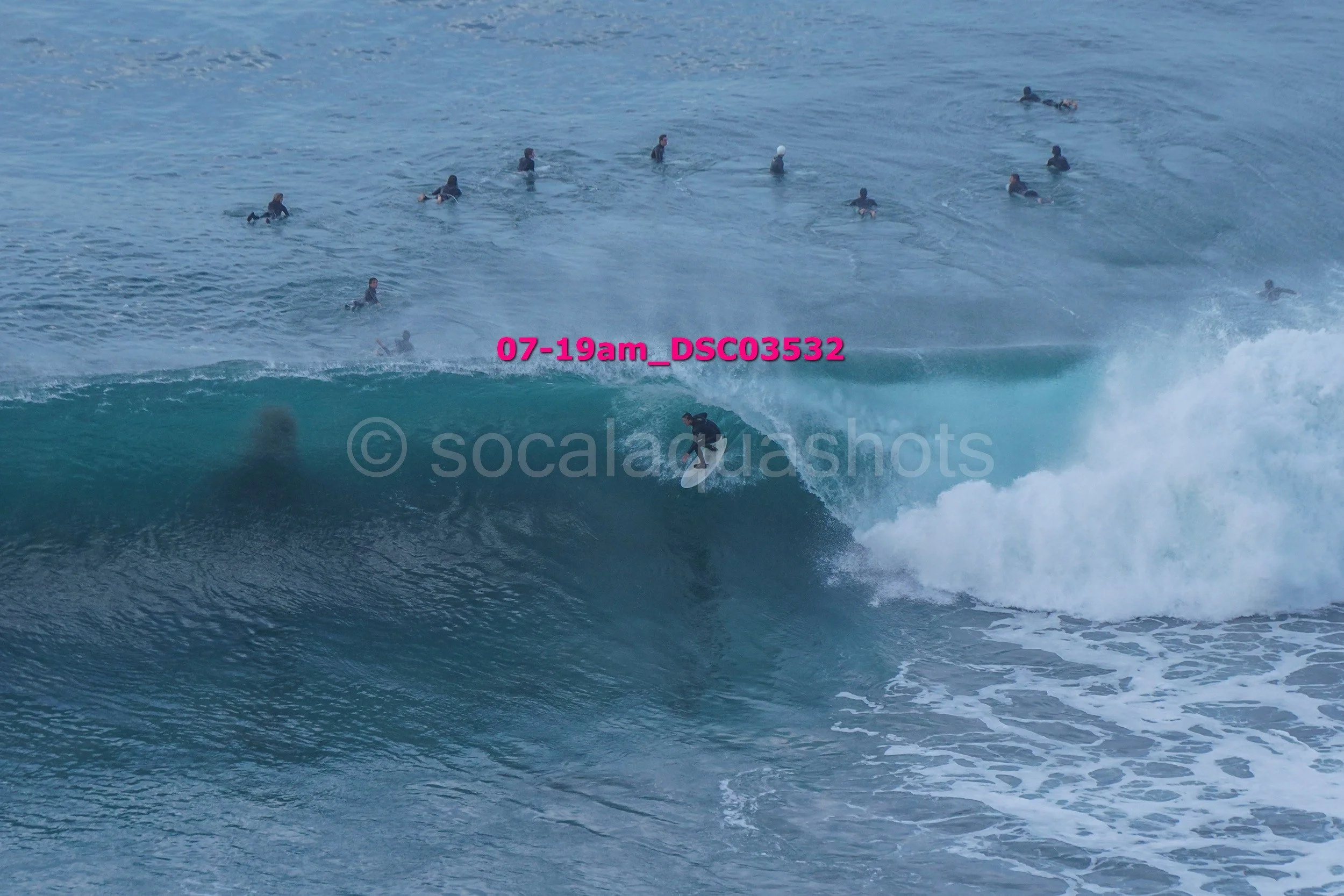 A surfer riding inside a large ocean wave while multiple surfers wait in the water behind him.