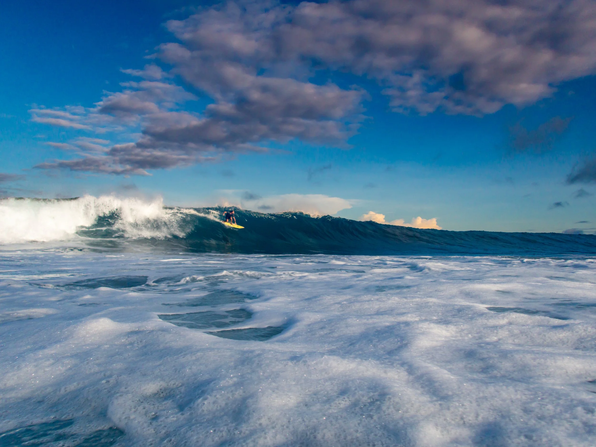 Surfer riding a large wave under a blue sky with clouds, foamy ocean in foreground.