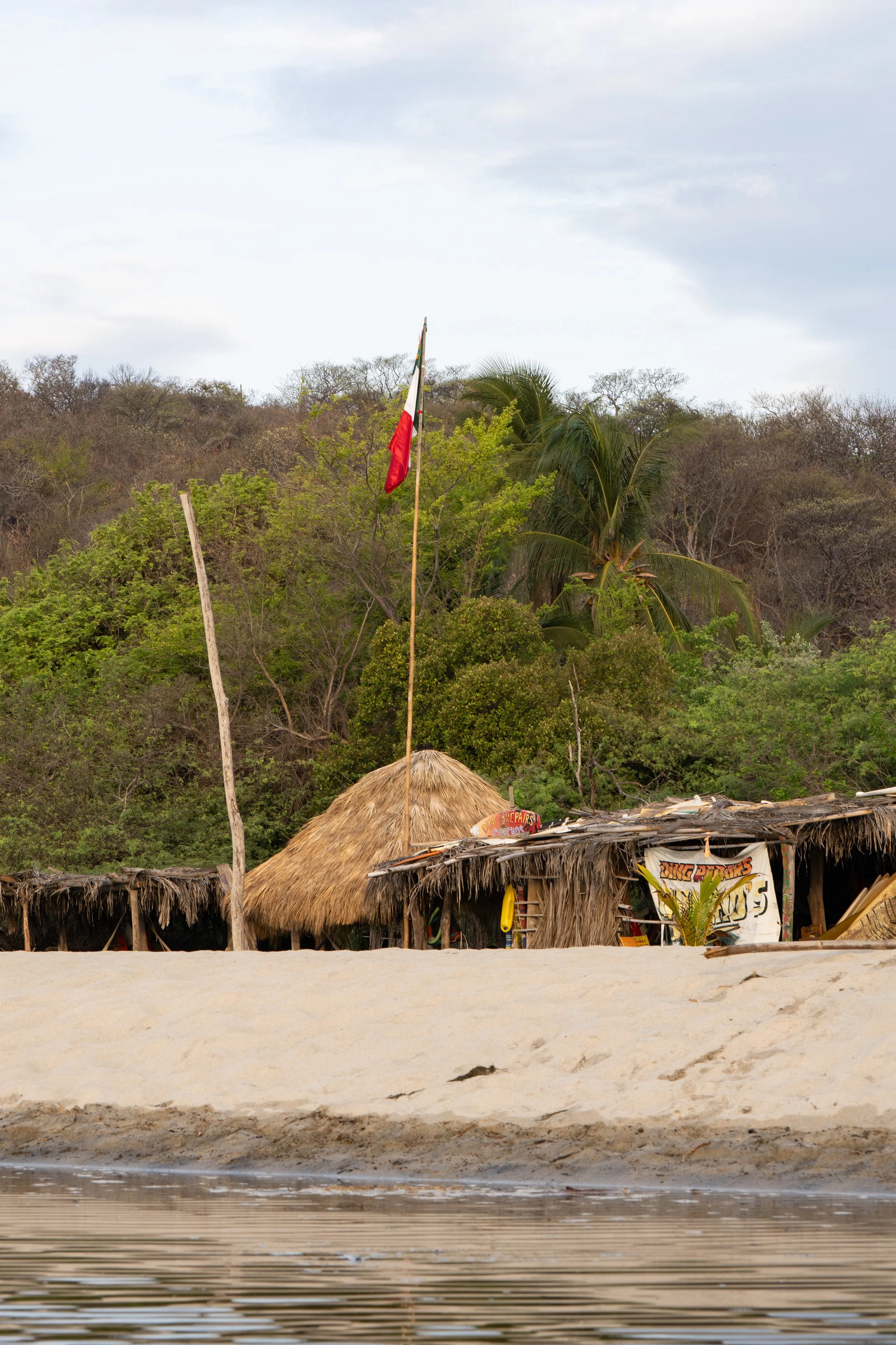 Beachfront huts with thatched roofs and a Mexican flag on a tall pole, set against a hillside with trees in the background, and water in the foreground.