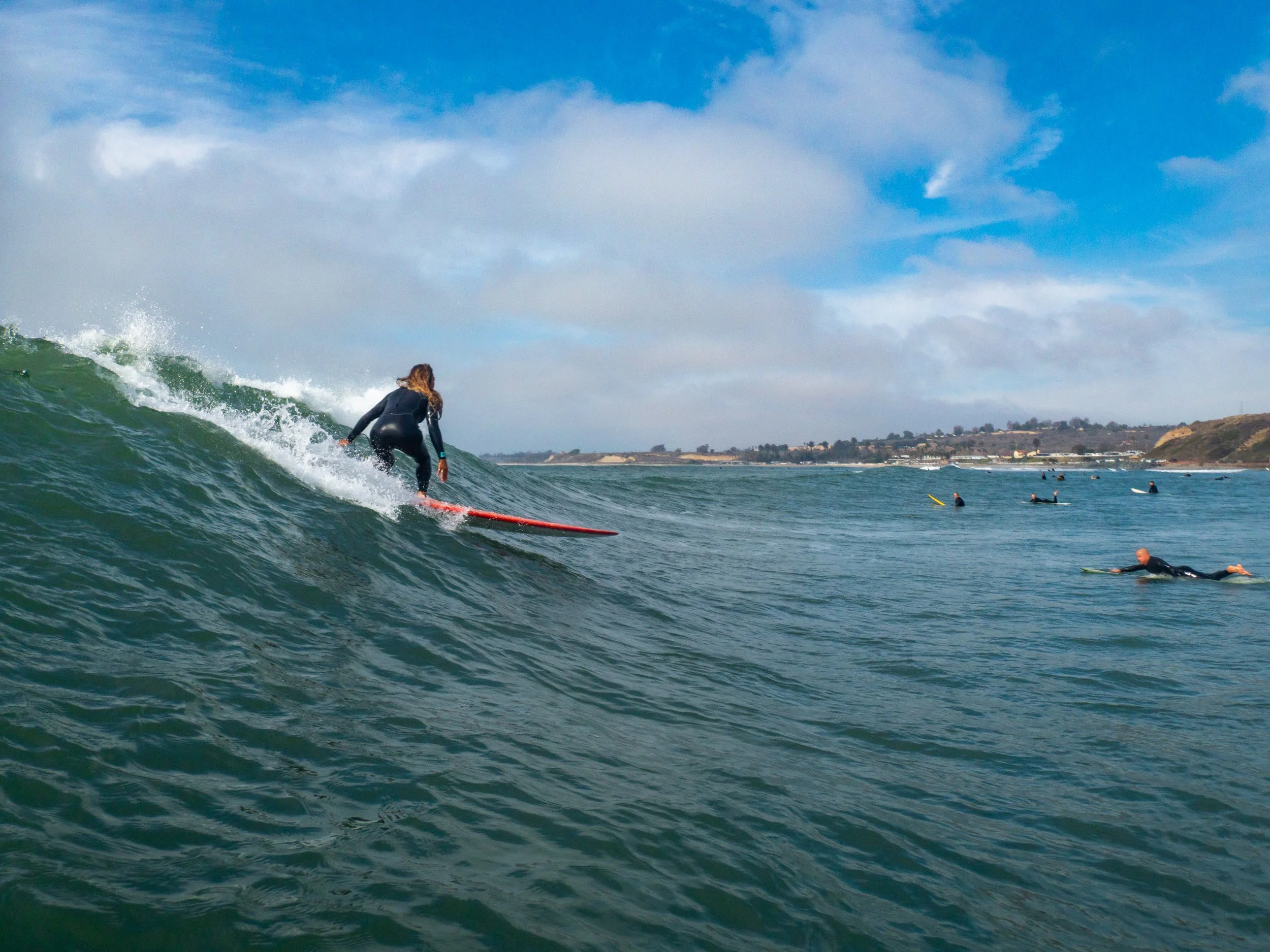 A female surfer riding a wave in the ocean with other surfers in the distance and a shoreline with buildings and hills under a partly cloudy sky.