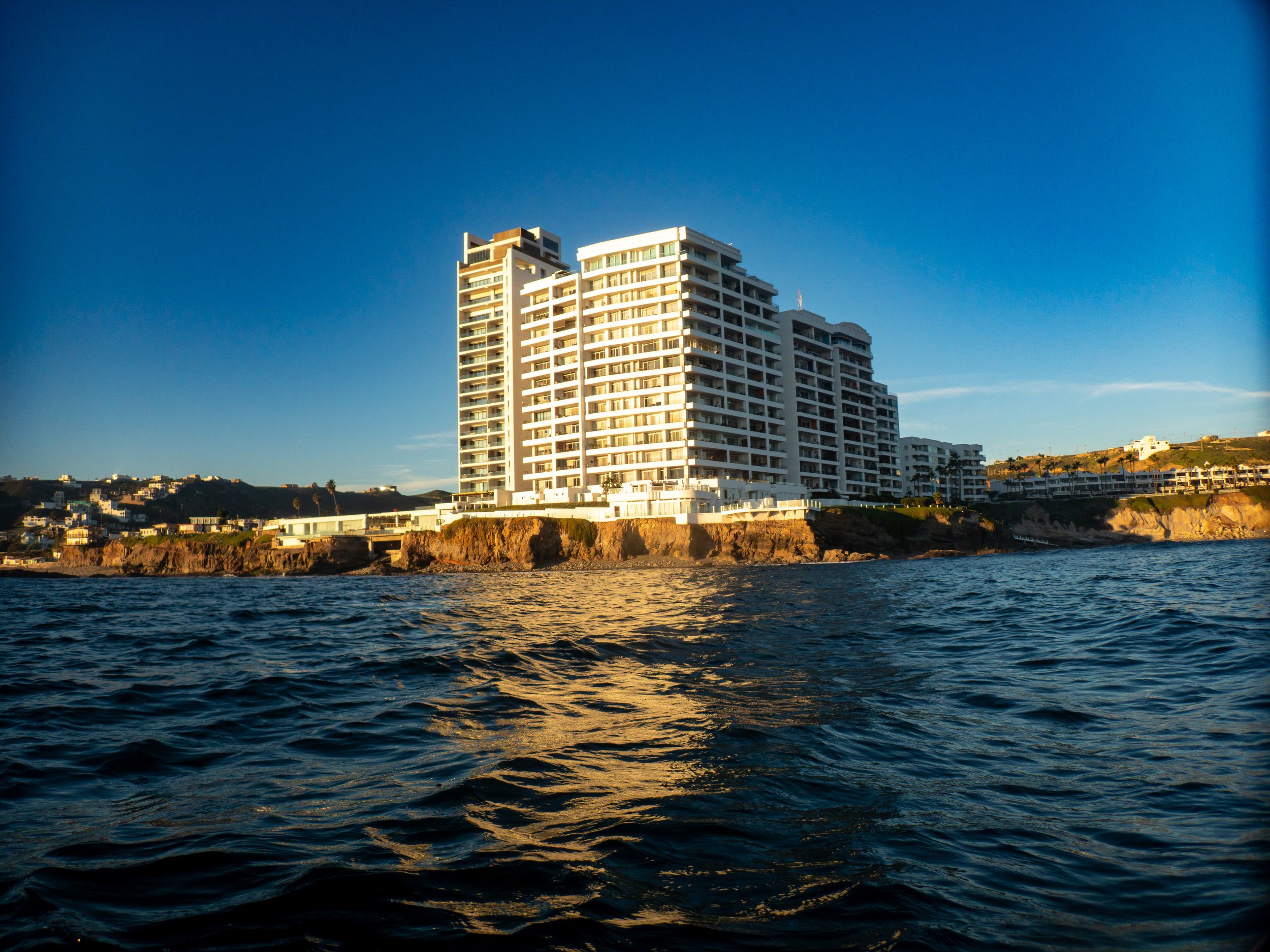High-rise white residential building on land next to ocean with waves and cliffs, clear blue sky.