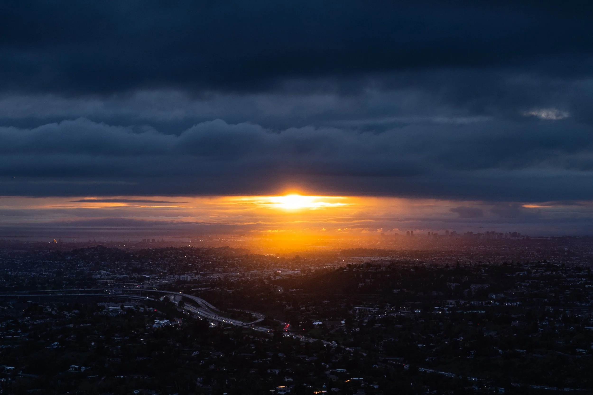 Sunset over a city skyline with dark clouds in the sky and a bright glow of the setting sun near the horizon.