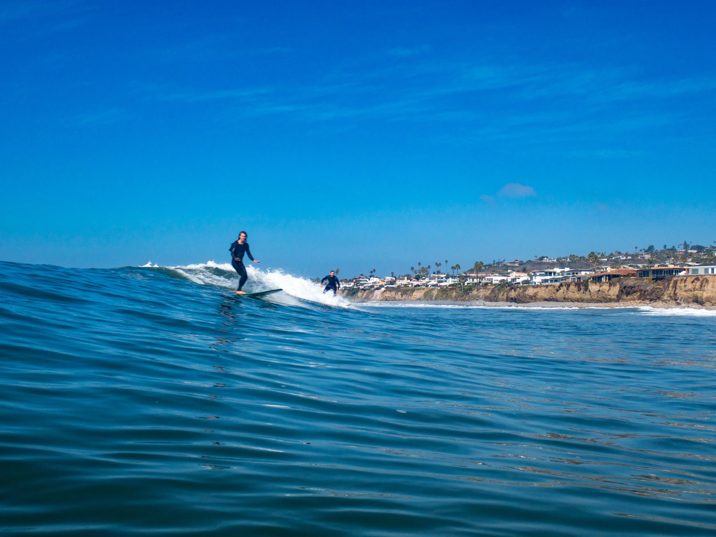 Two people surfing on a blue ocean with a coastal town and palm trees in the background under a clear blue sky.