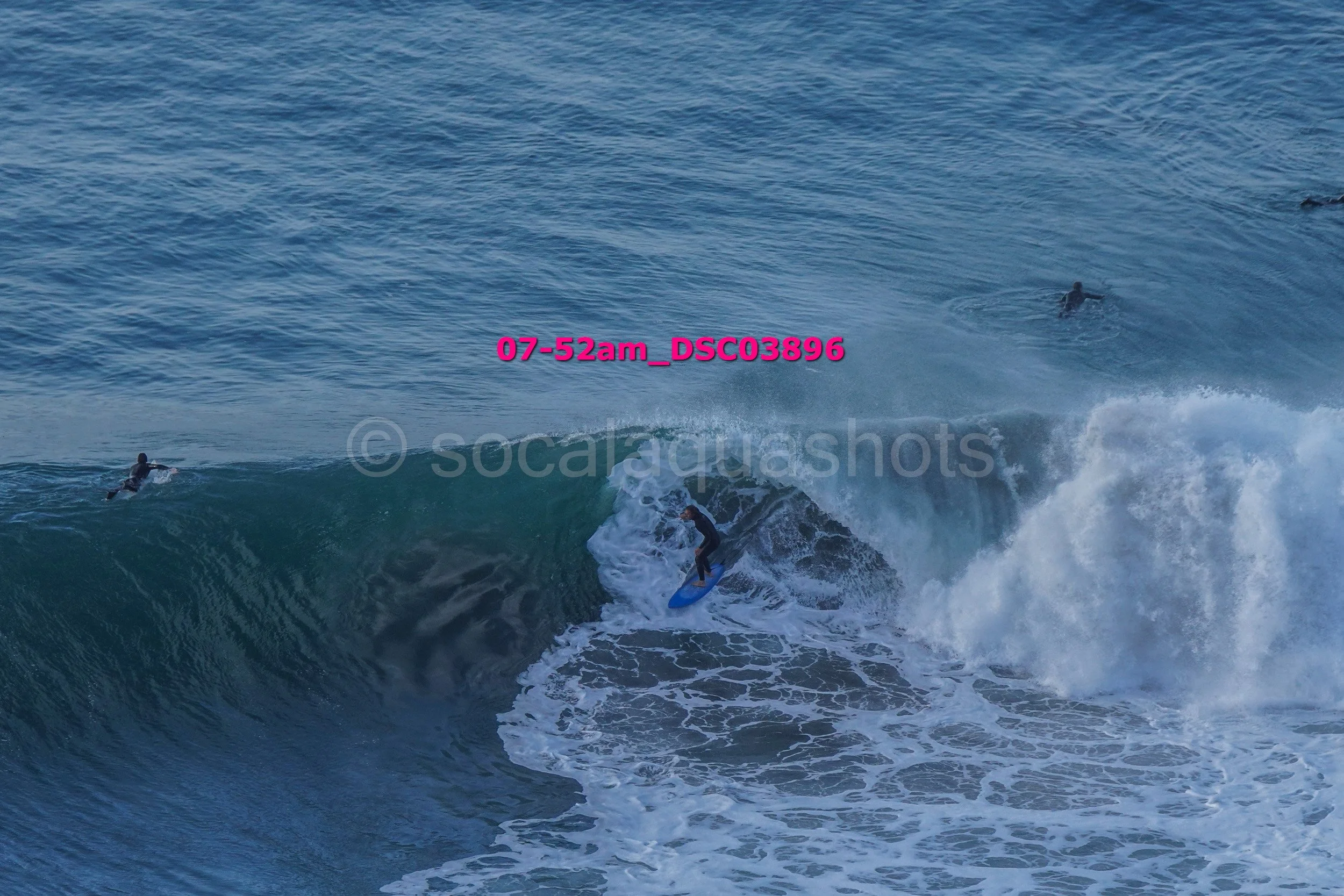 A person surfing on a wave in the ocean with two other surfers swimming nearby.