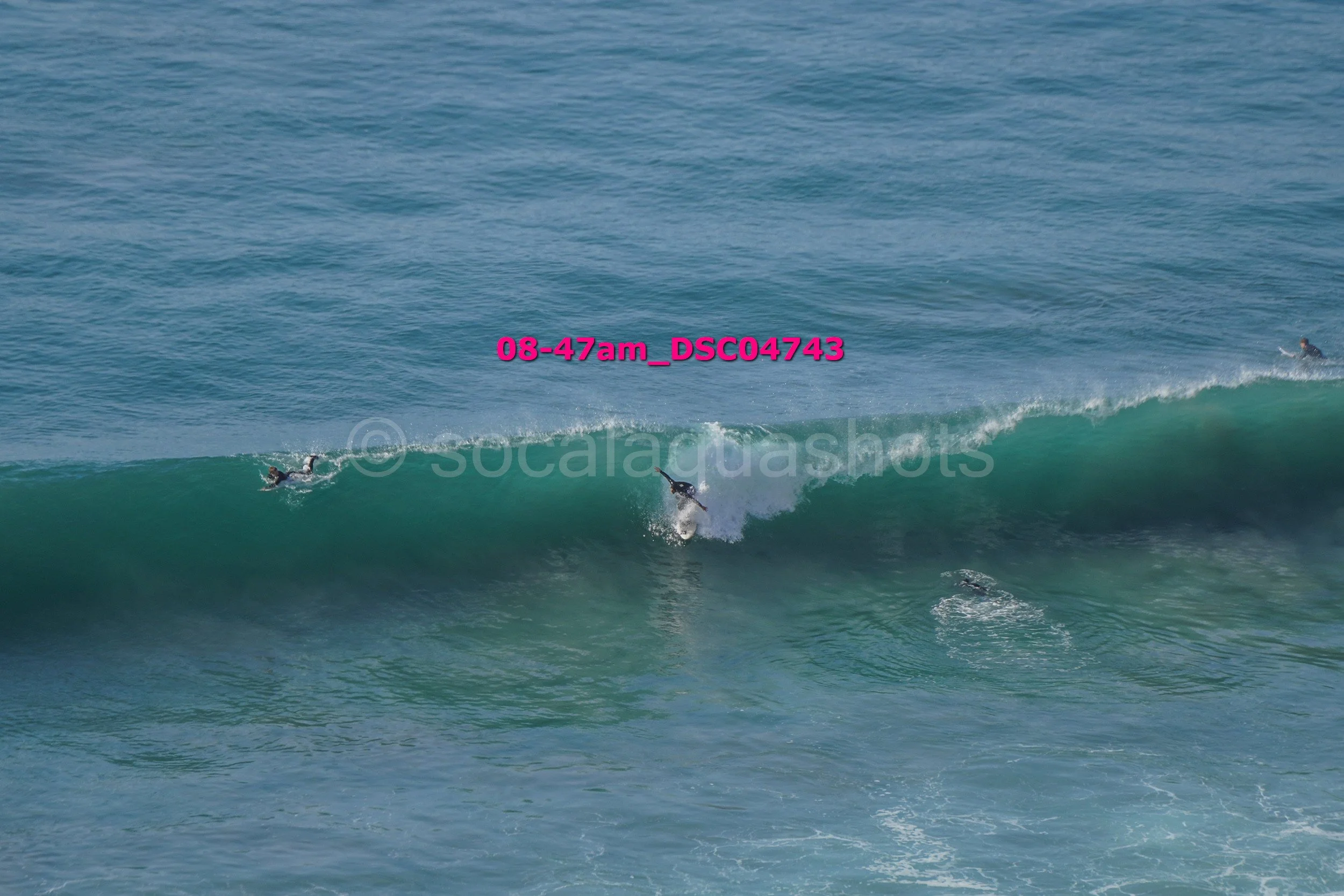 Three surfers riding a large ocean wave, one in the center and two on the right, with the water in shades of blue and green.