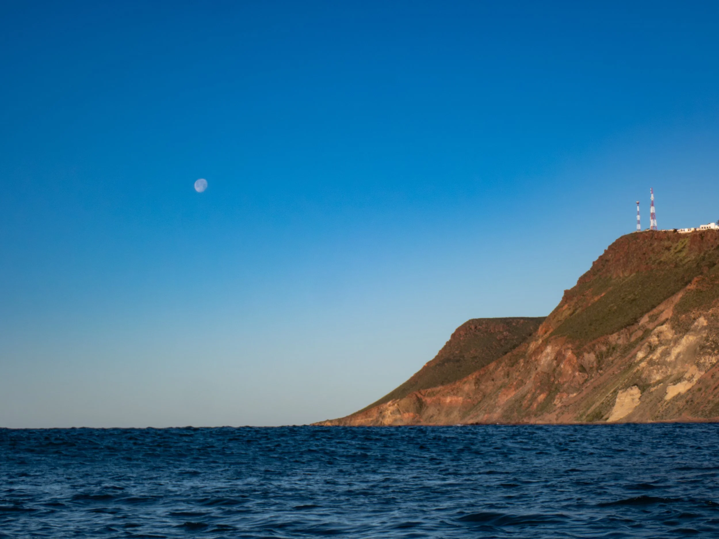 View of ocean with a steep hill or mountain in the background, topped with communication towers, under a bright blue sky with the moon visible.