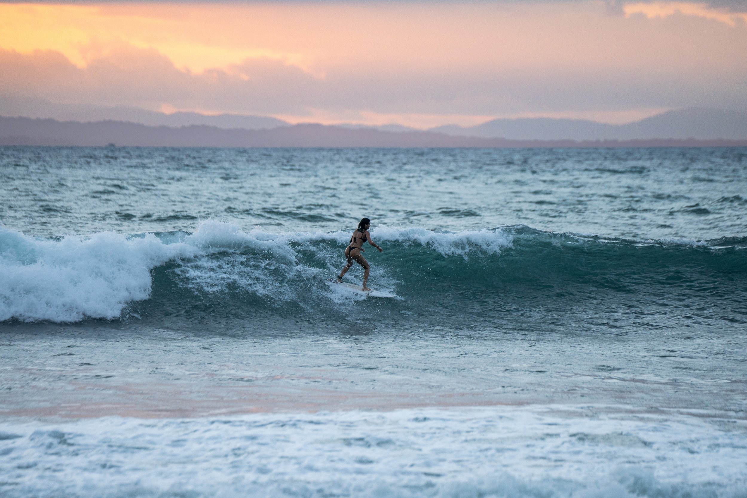 A person surfing on a wave in the ocean during sunset with mountains in the background.