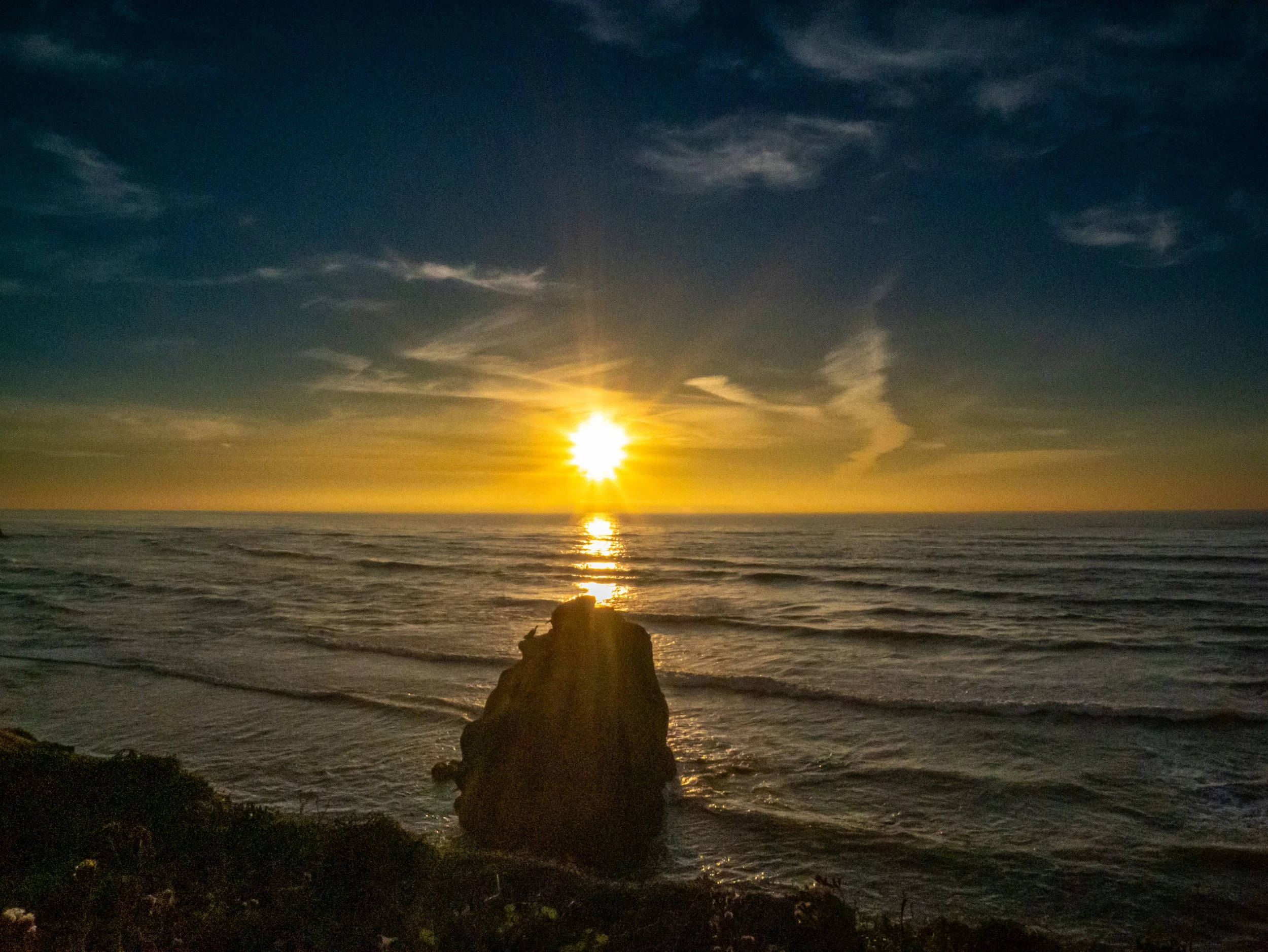 Sunset over the ocean with a large rock in the foreground and clouds in the sky.