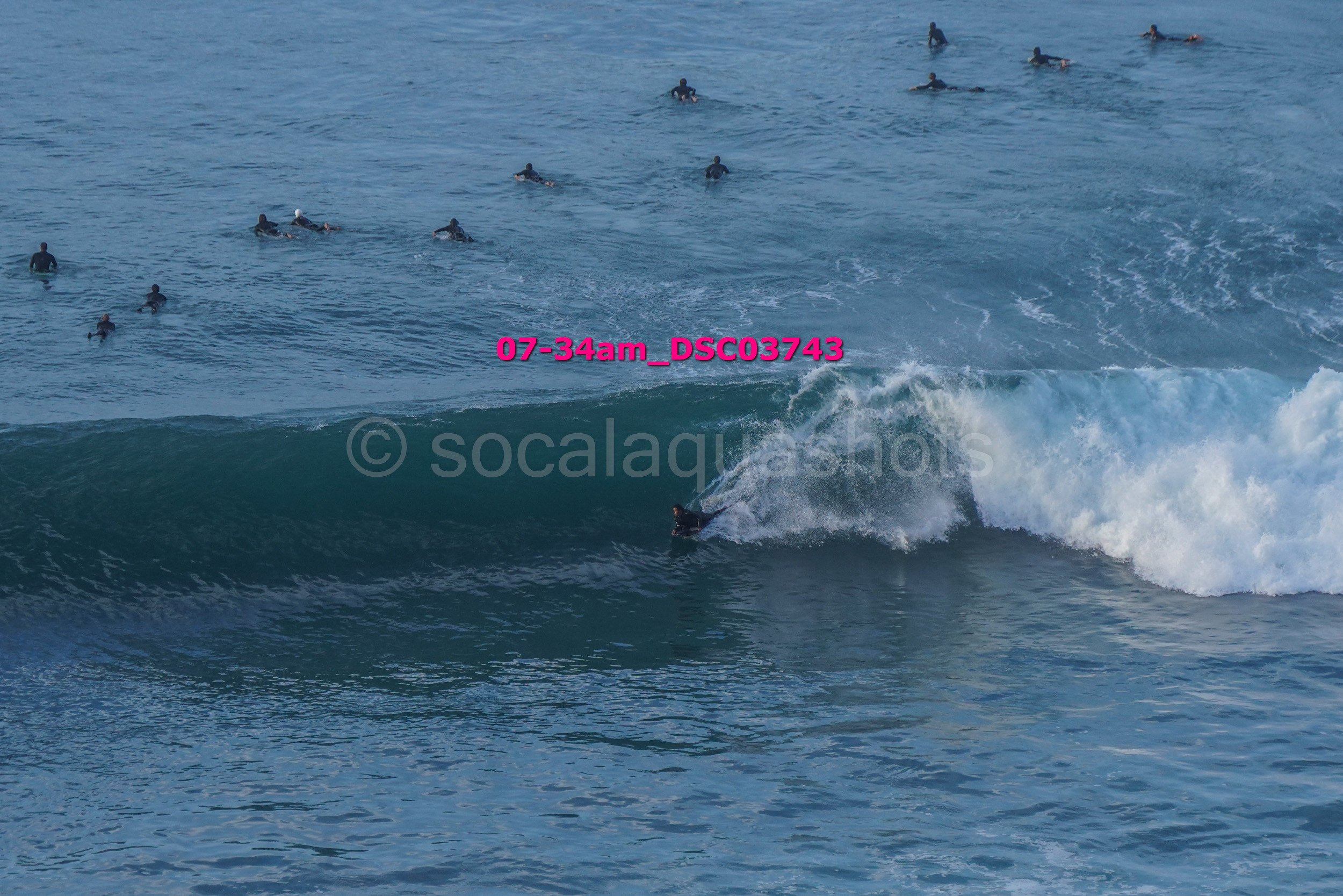 Surfer riding a wave with numerous people in the water and some on surfboards in the background.