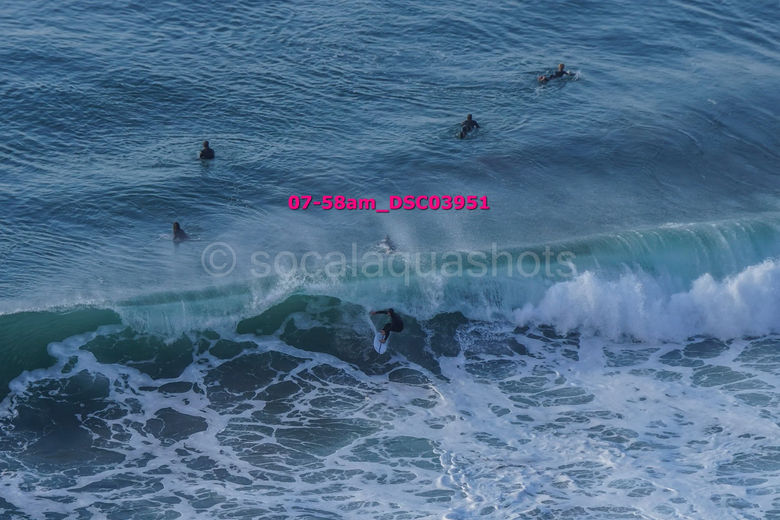 A surfer riding a wave in the ocean with several people swimming or surfing in the background.