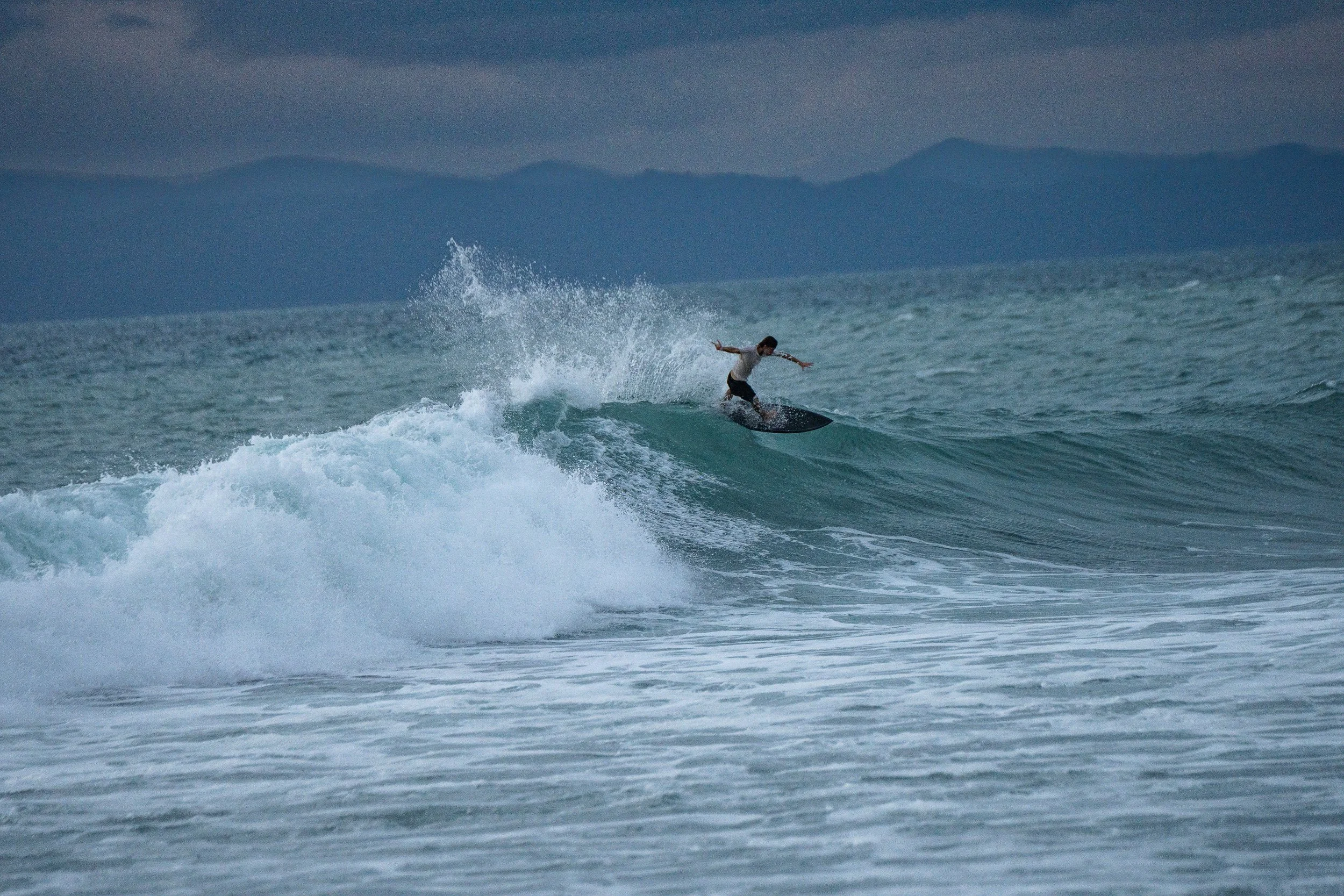 A person surfing on a wave in the ocean with mountains visible in the background during cloudy weather.