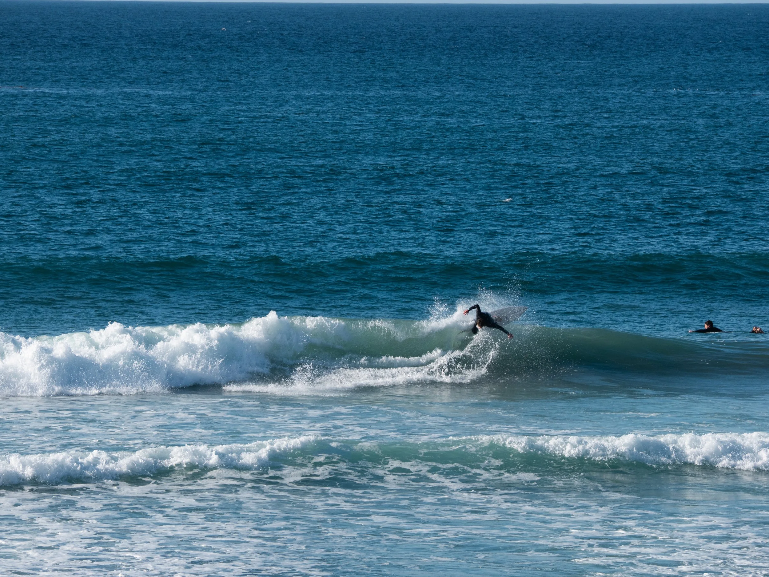 A person surfing on a wave in the ocean, with other surfers nearby, under clear blue skies.