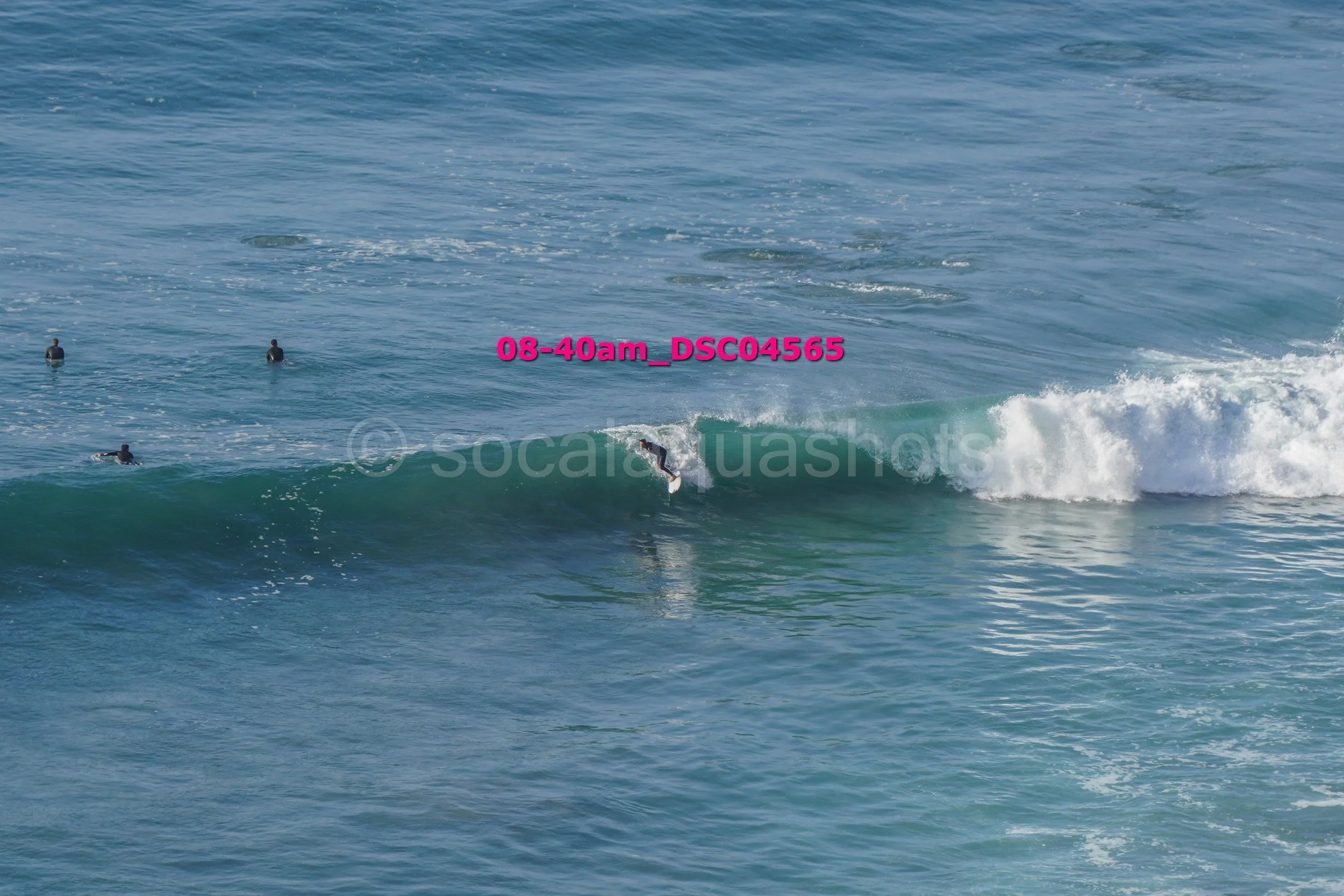 Surfer riding a wave with three surfers in the water and white surf to the right.