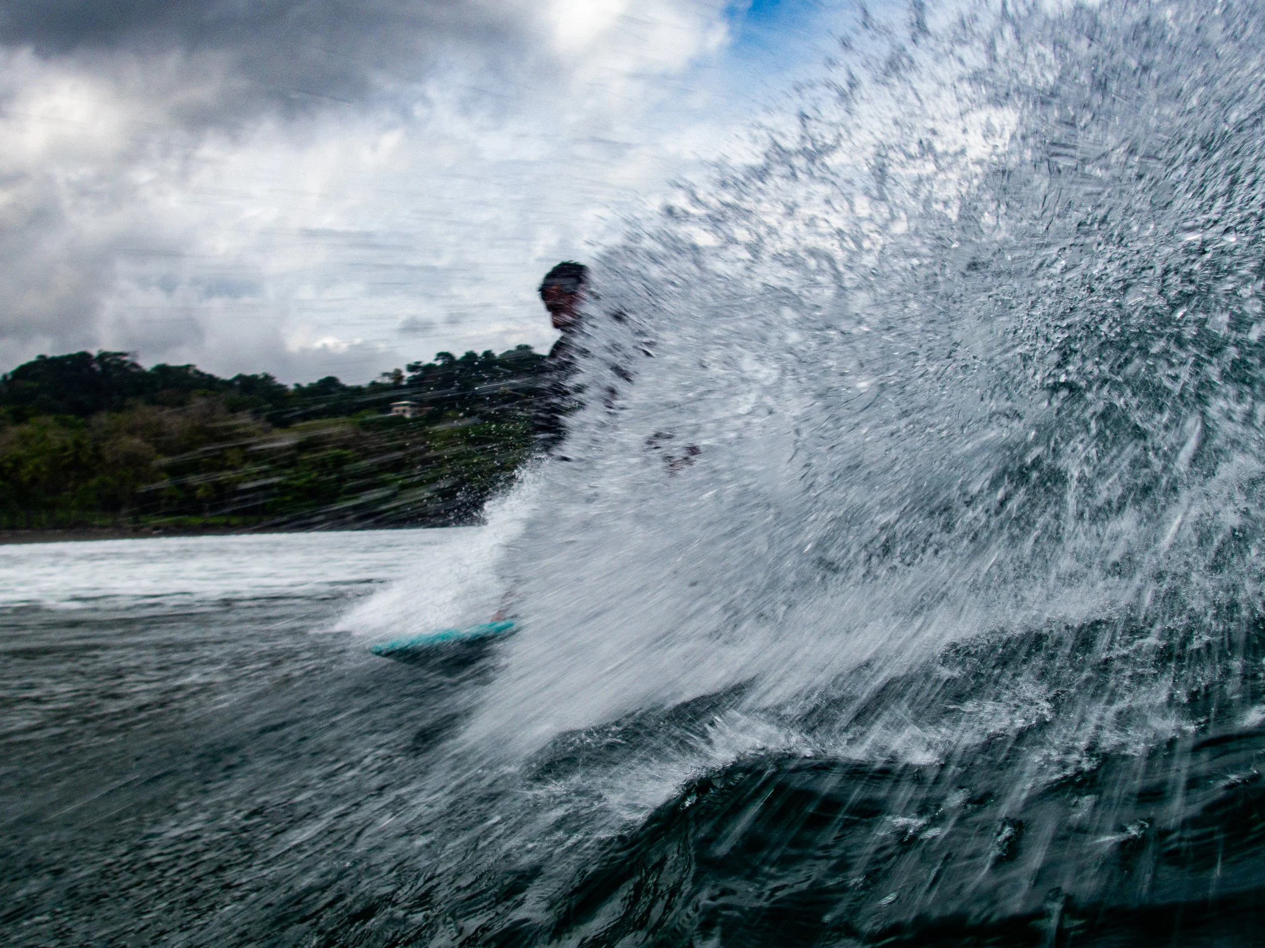 Surfer riding a wave, water splash obscuring view, cloudy sky, coastal landscape in background.