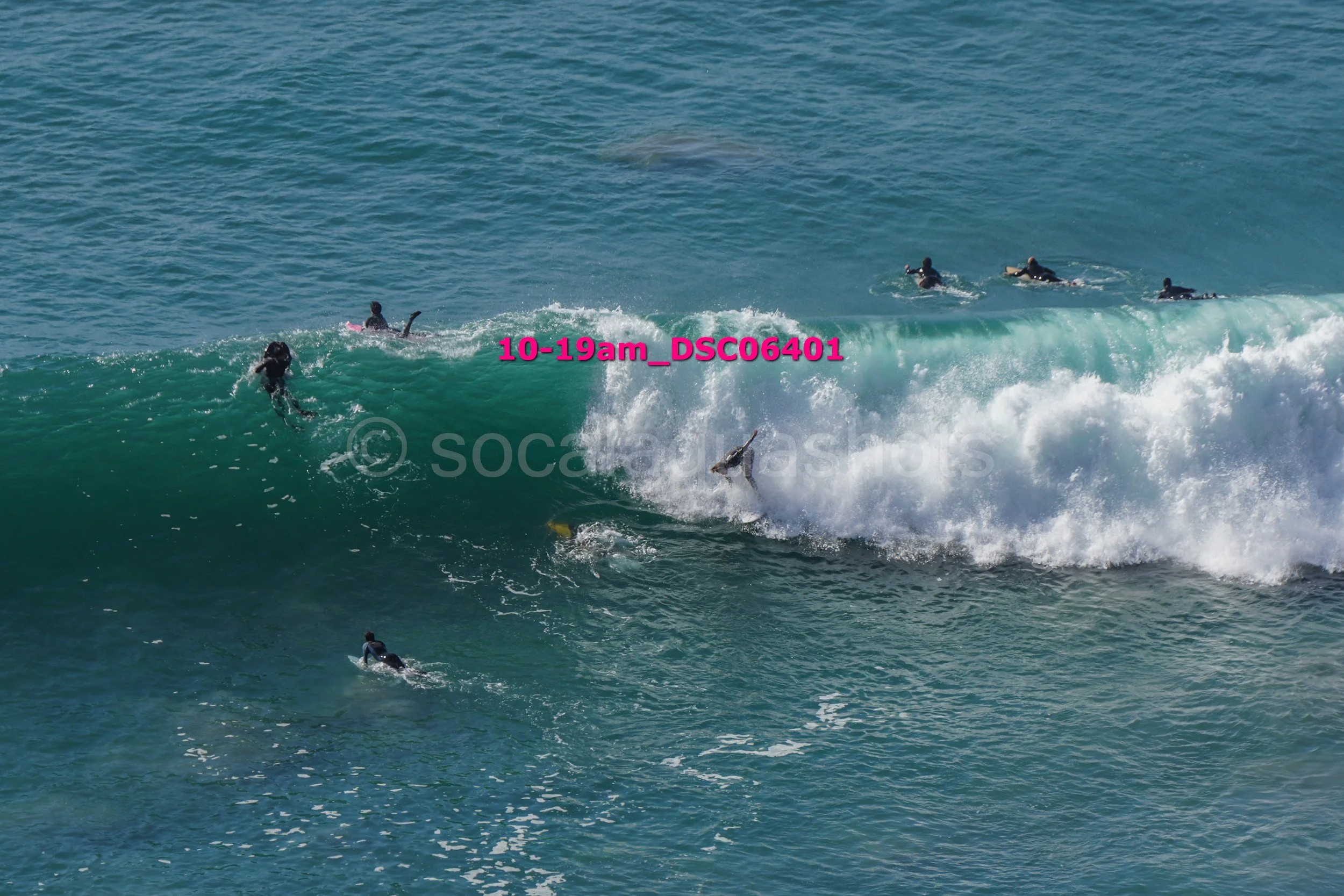 Surfers riding and paddling on ocean waves with people in the background.