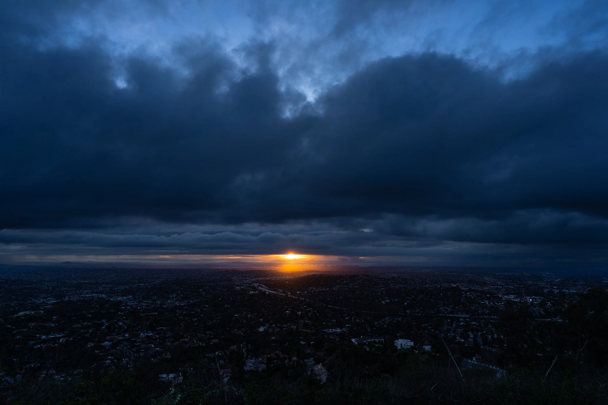 Dark, cloudy sky over a city at sunset with the sun near the horizon, casting a glow through the clouds.