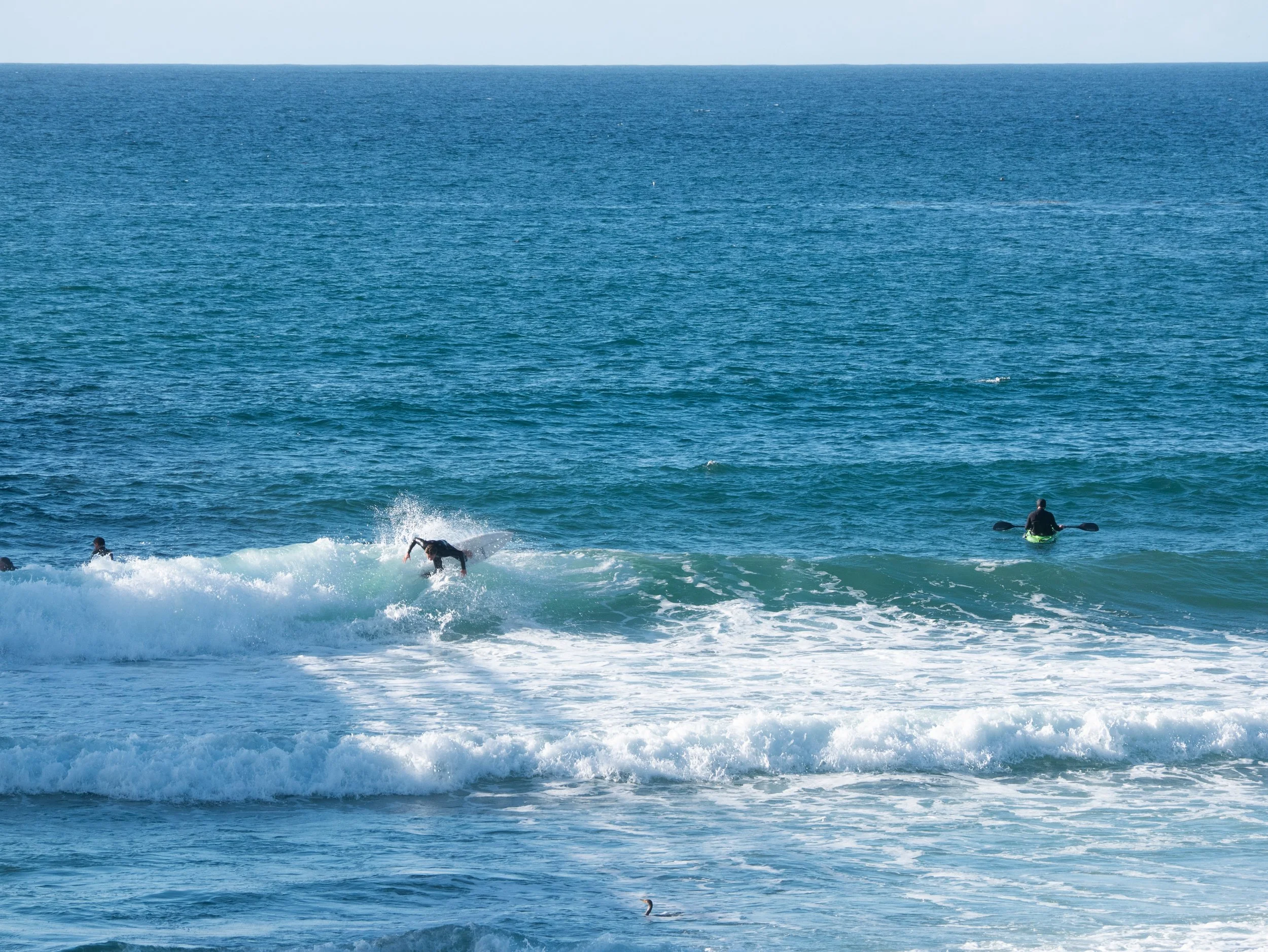 People surfing and paddling in the ocean with waves, under a clear blue sky.