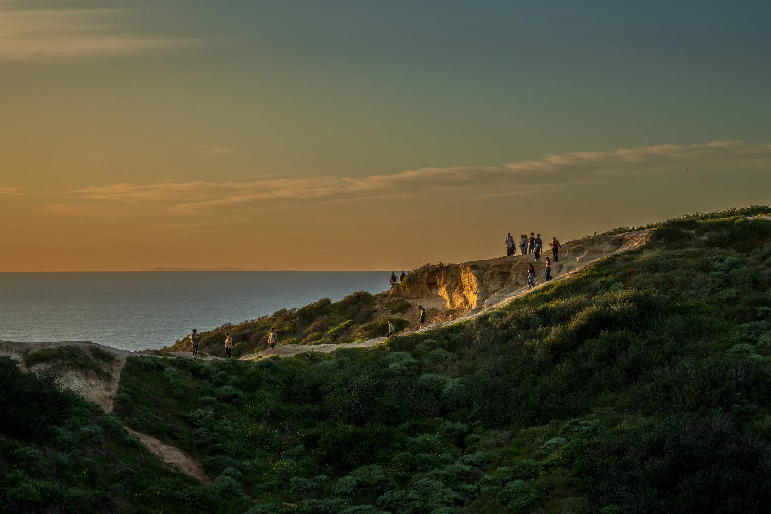 People hiking on a hillside overlooking the ocean during sunset.