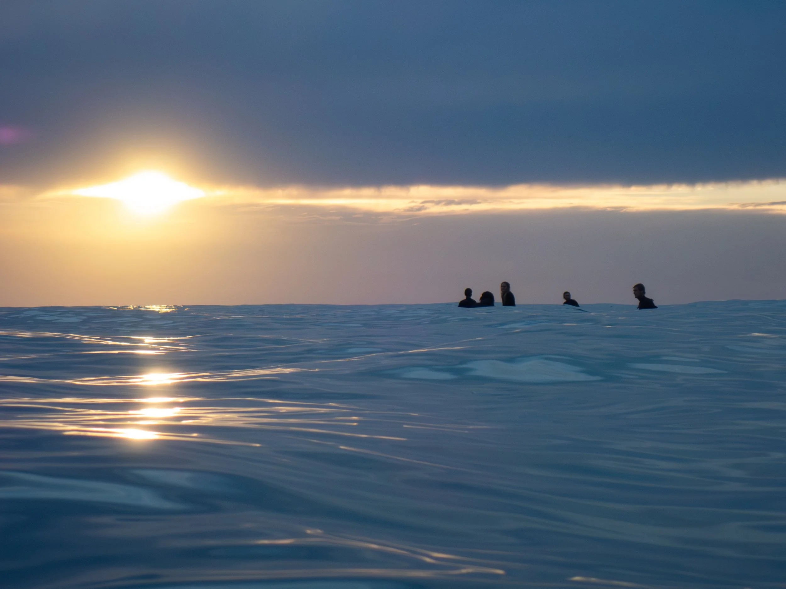 Silhouettes of people swimming in the ocean at sunset, with dark clouds and golden sunlight reflecting on the water.