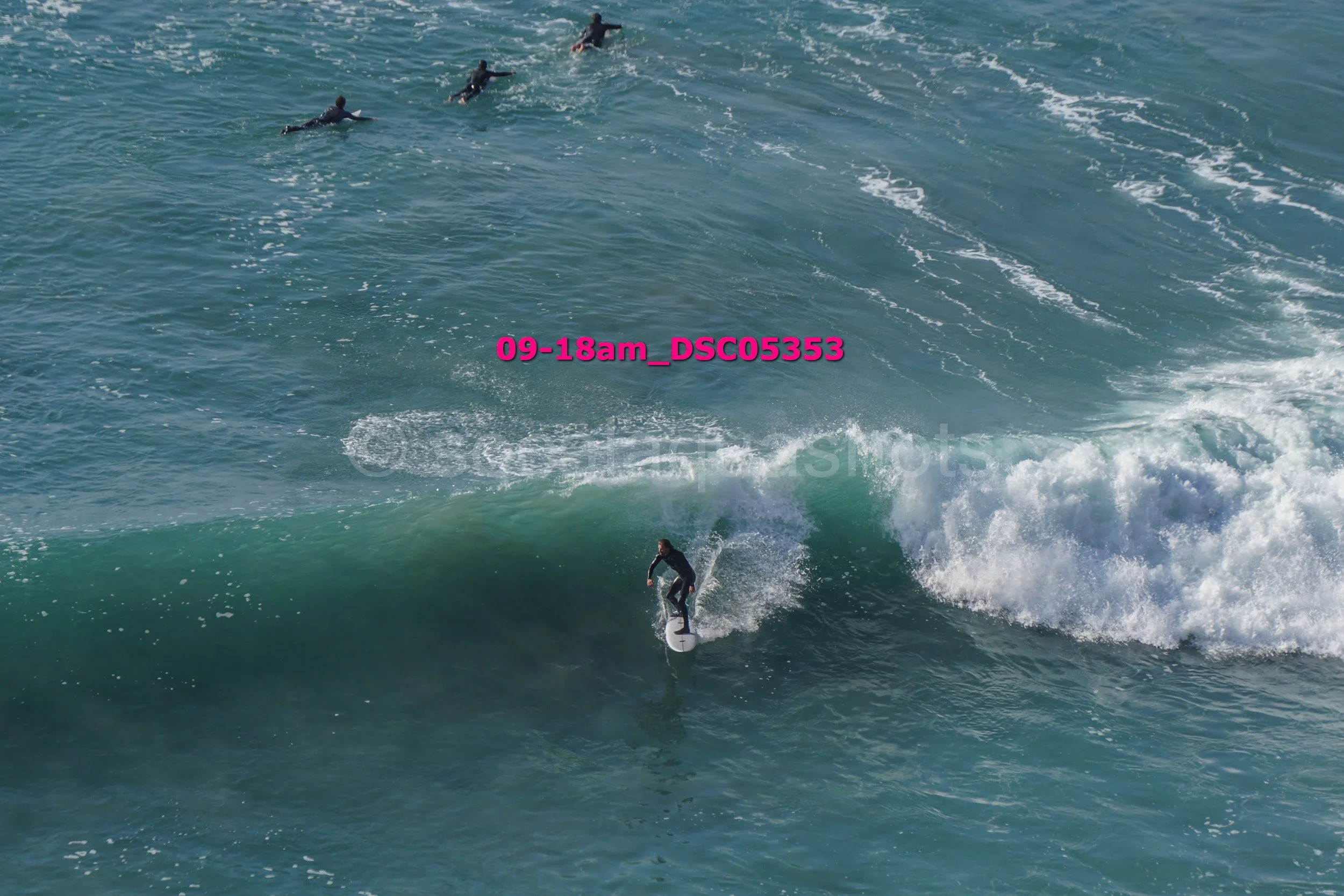 A person surfing on a large ocean wave with three other surfers in the distance.