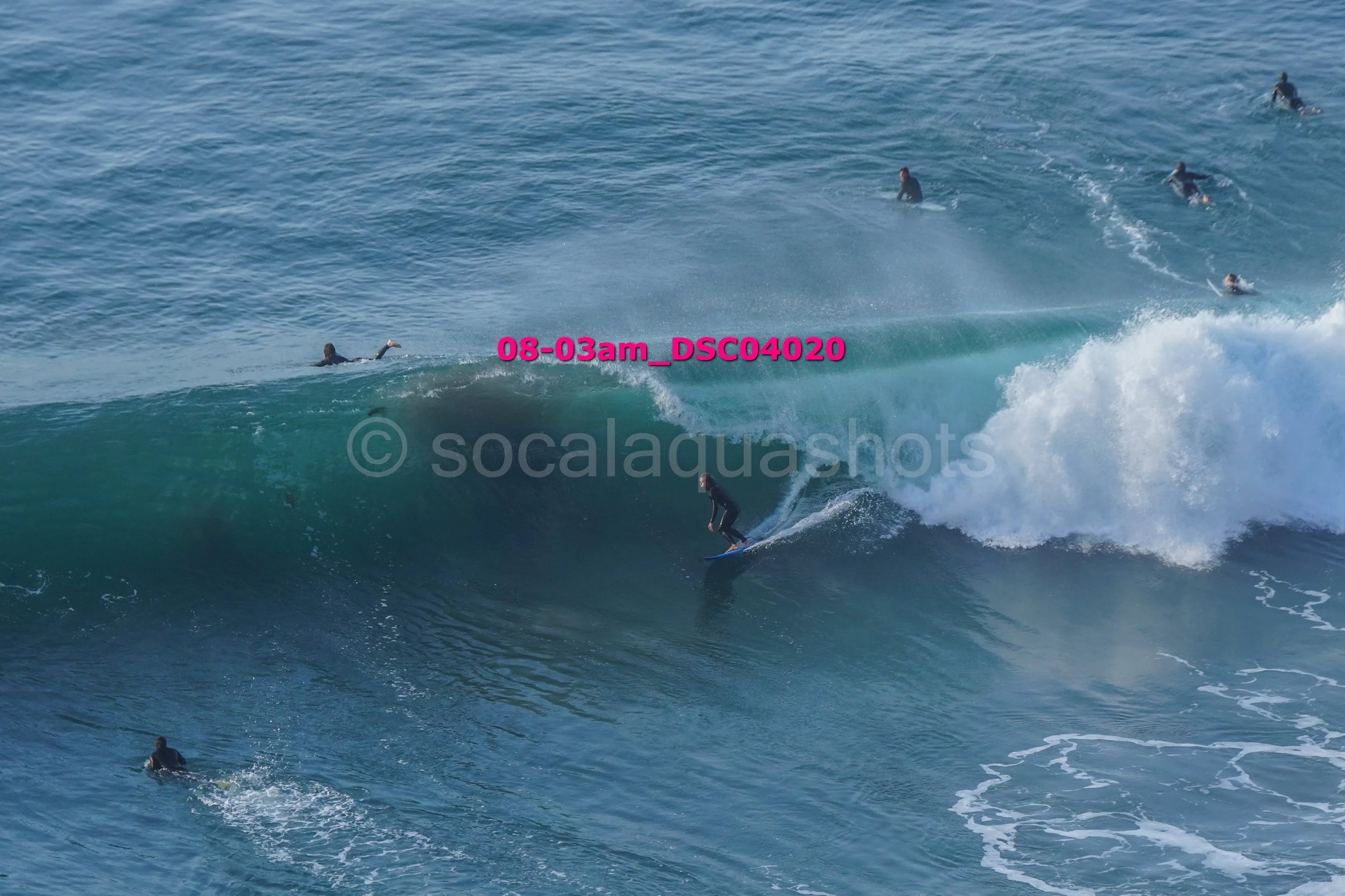 A surfer in black wetsuit riding a large ocean wave with multiple surfers in the water nearby.