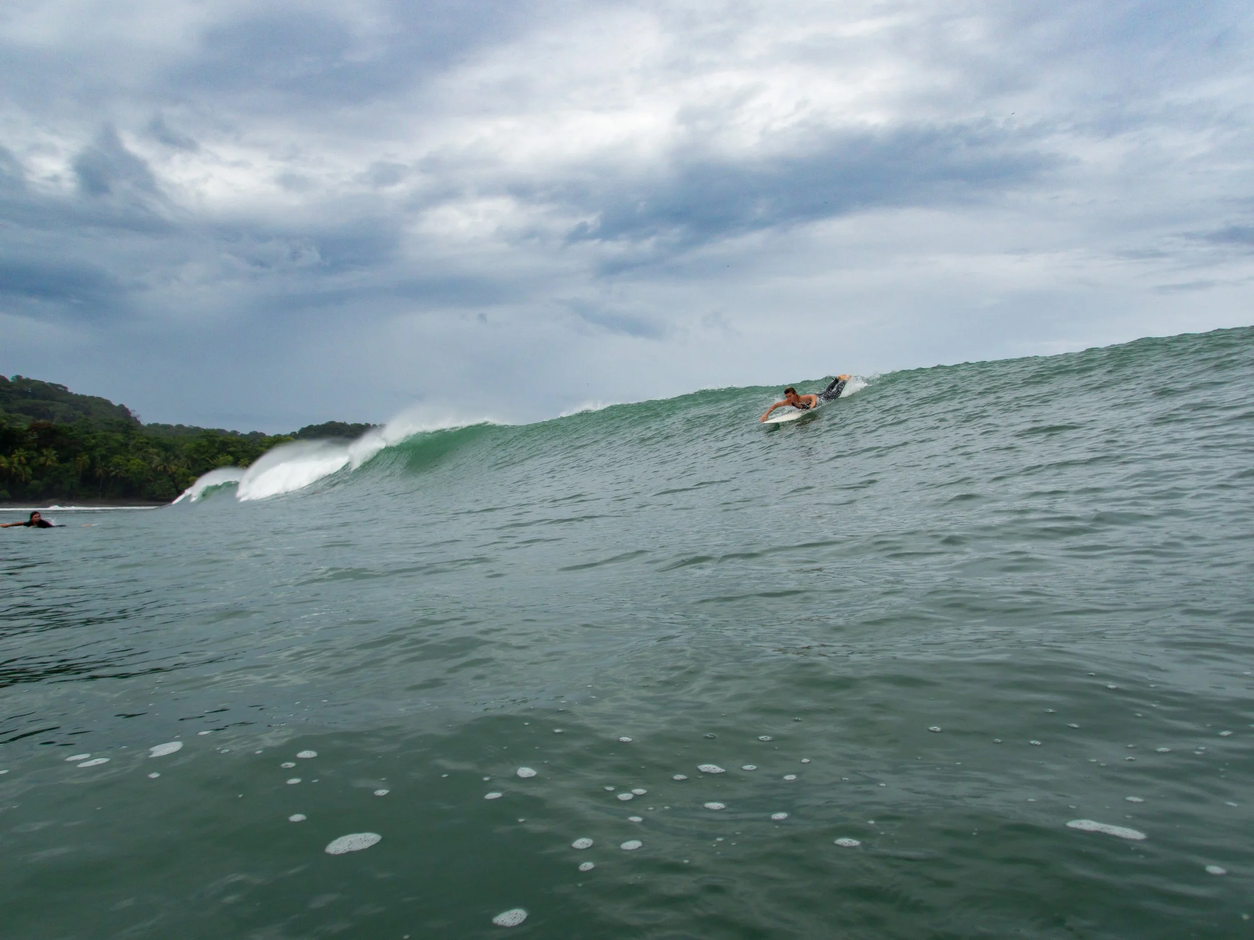 Surfer riding a large wave in the ocean with a cloudy sky and shoreline in the background.