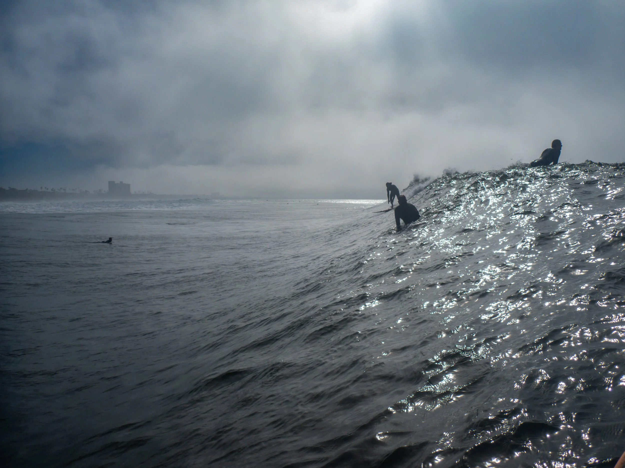 Several surfers are riding a wave at the beach under cloudy sky, with one surfer in the water and others on the wave.