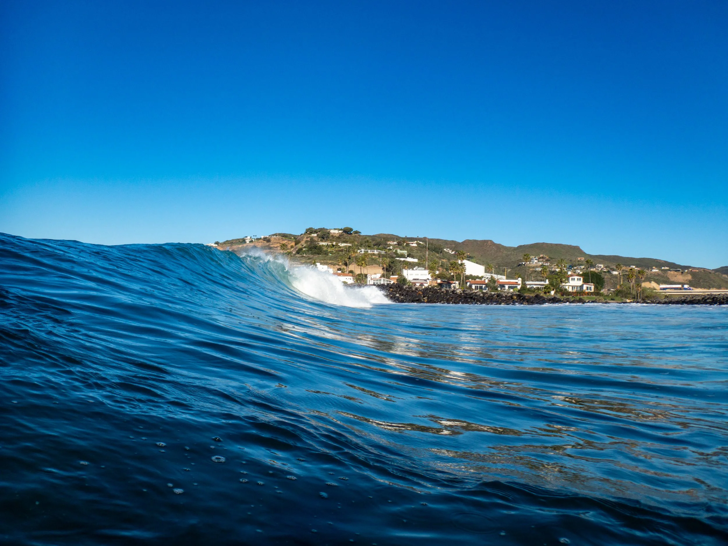 A clear blue ocean wave approaching the shoreline with houses and rolling hills in the background under a bright blue sky.
