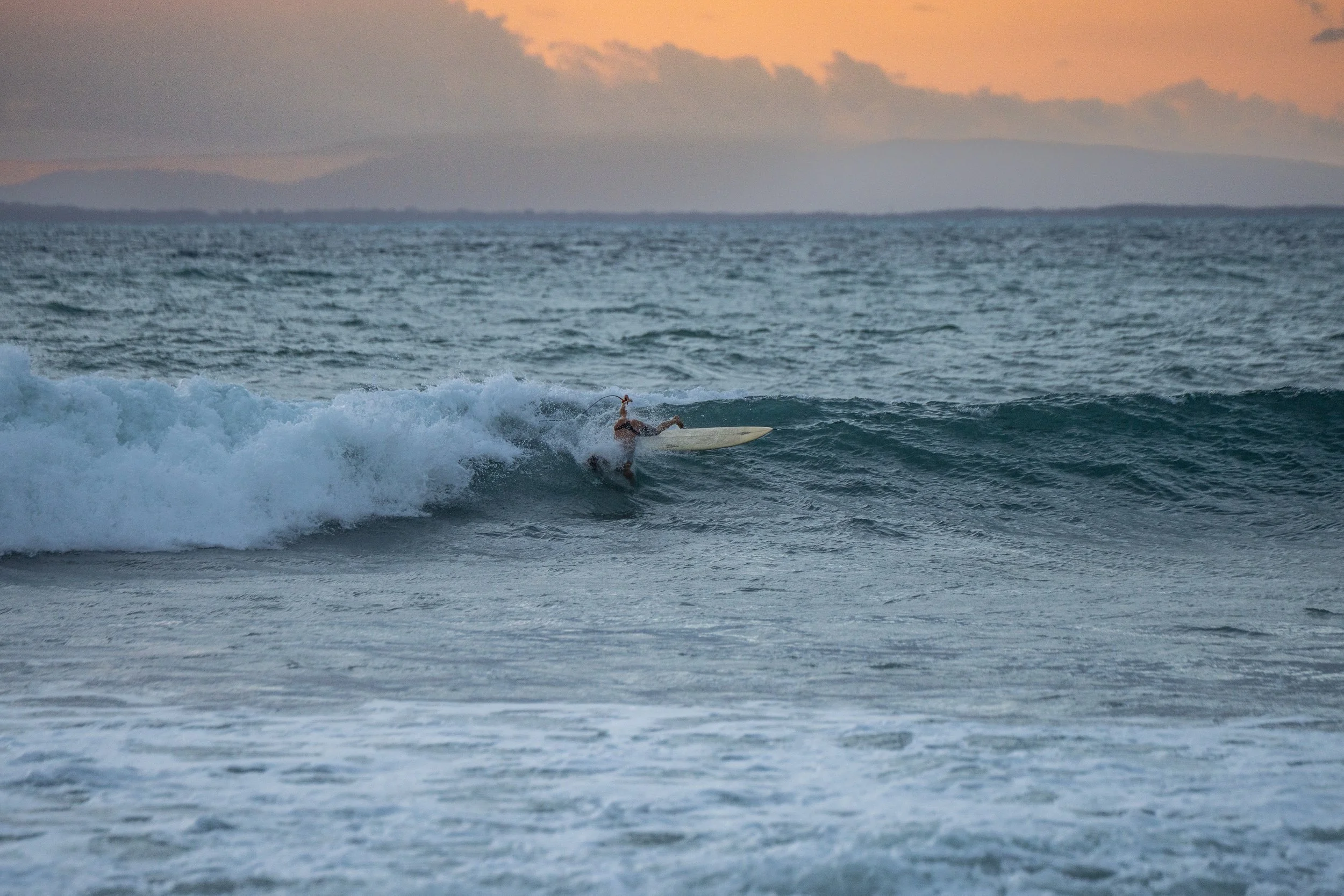 A surfer riding a wave in the ocean at sunset with mountains in the background.