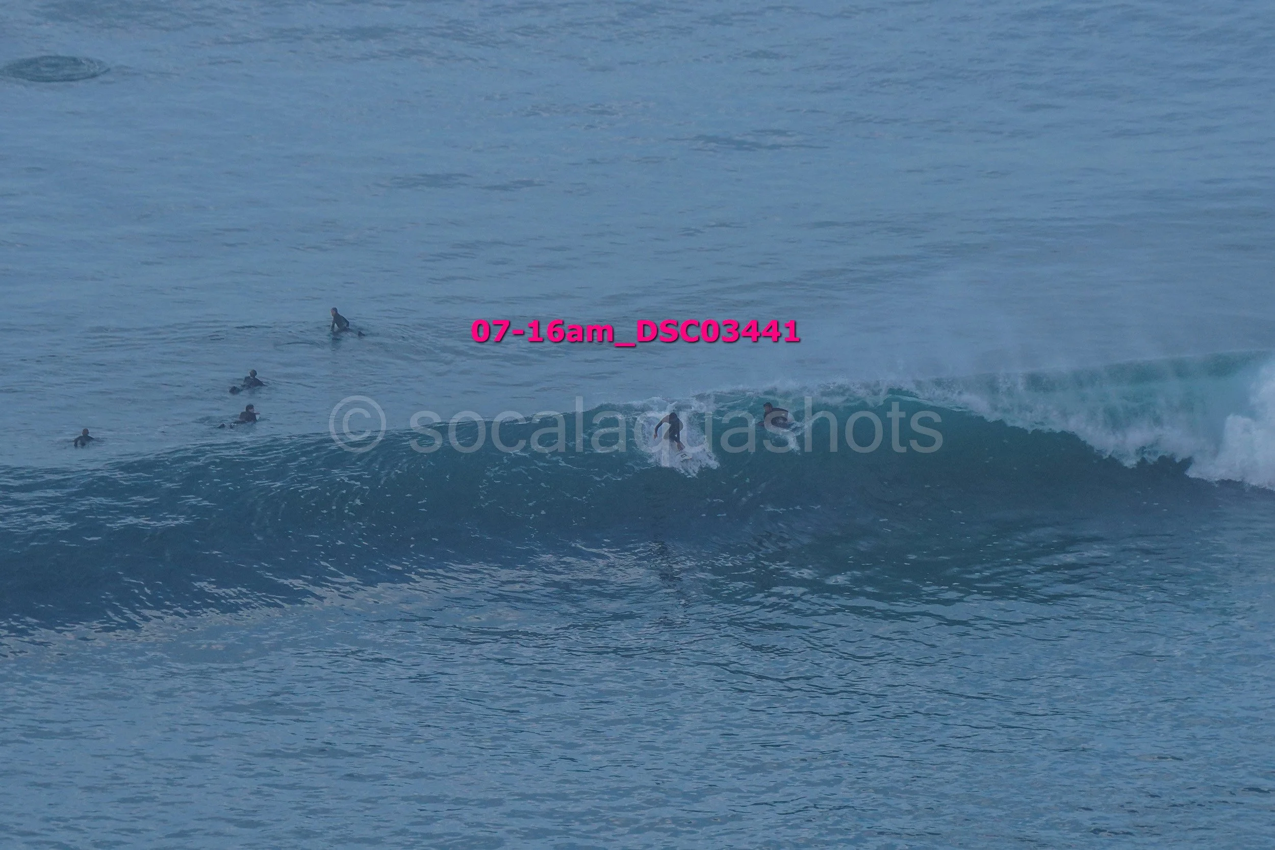 Surfer riding a wave in the ocean, while several other surfers wait in the water for their turn.
