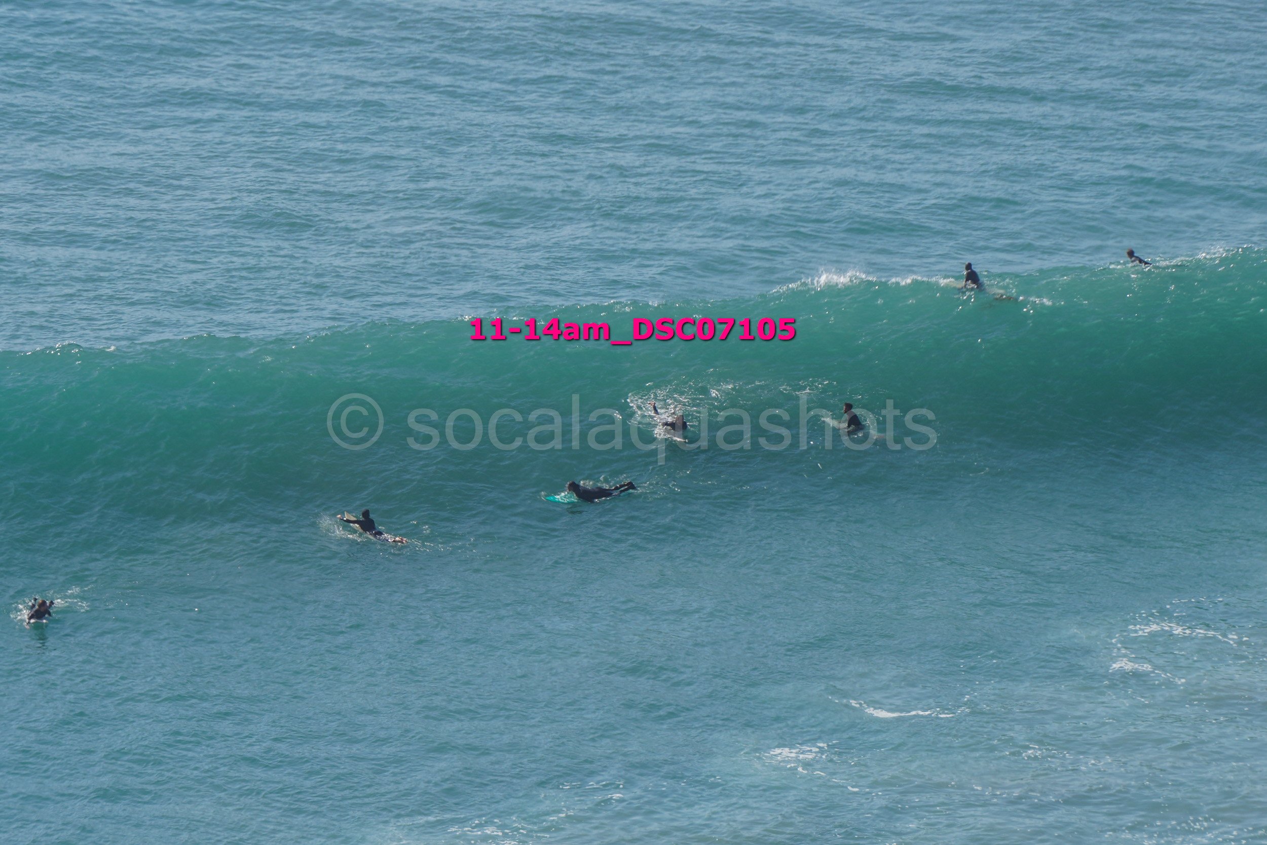 Several surfers riding and paddling on ocean waves with a few more in the distance, ocean water, and sky visible.