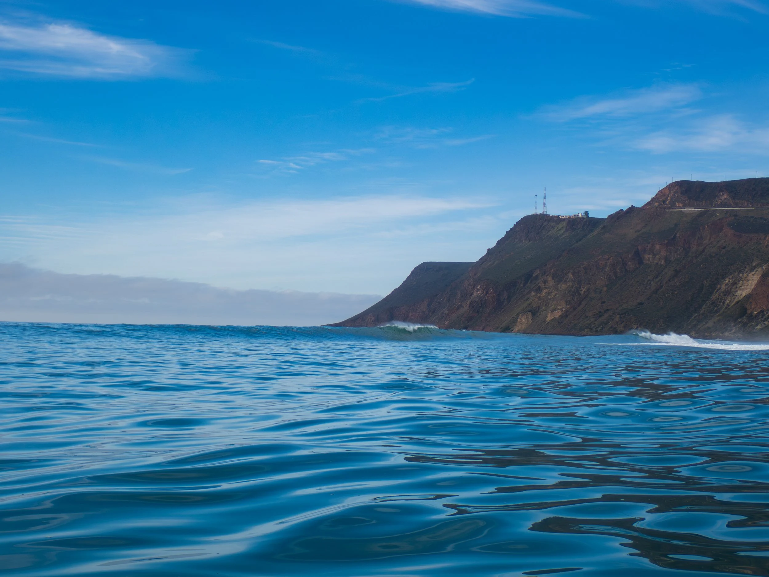 Ocean waves approaching a hillside with communication towers, under a partly cloudy sky.