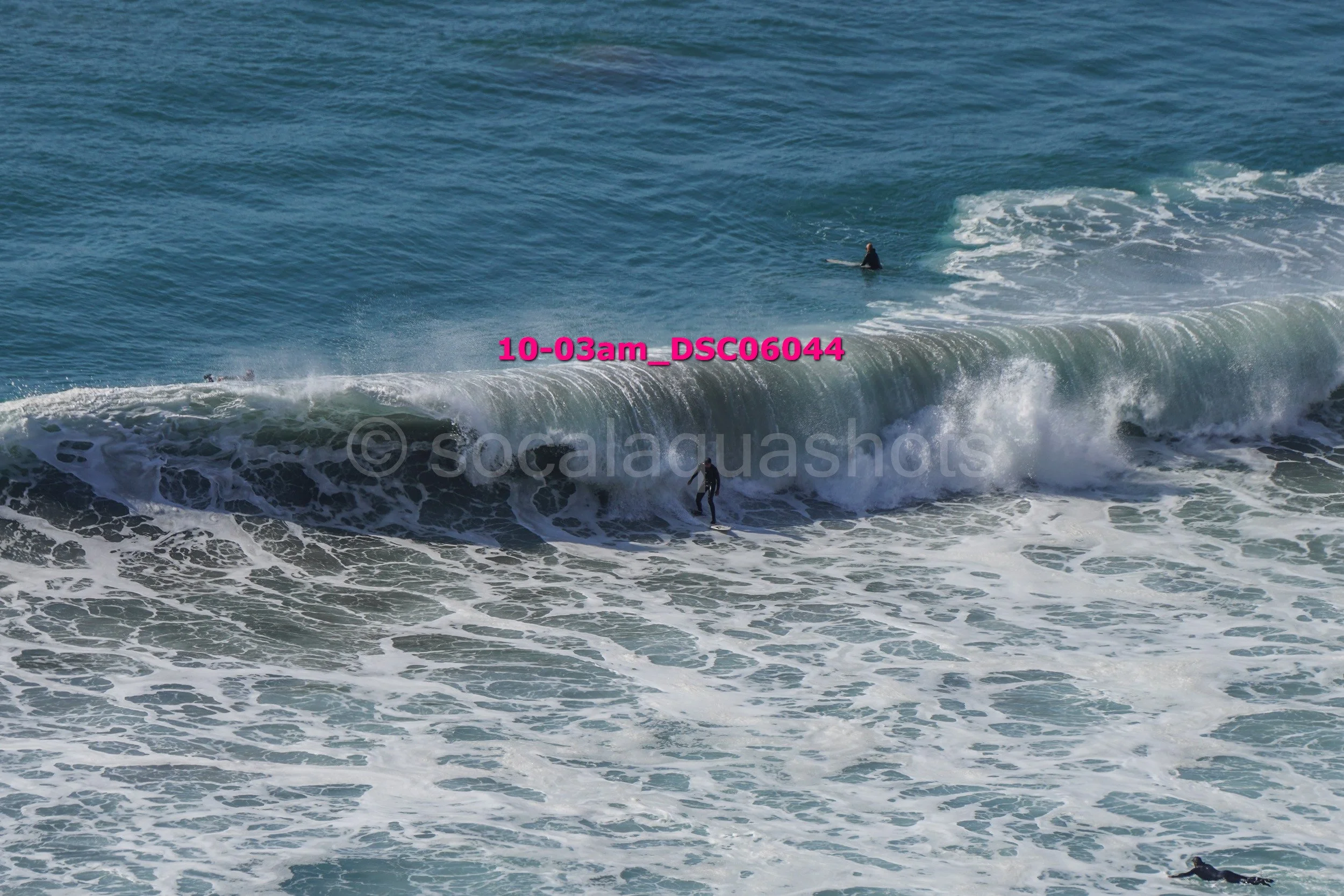 A surfer riding a large ocean wave with two other surfers in the water nearby, the water is blue with white foam and spray.