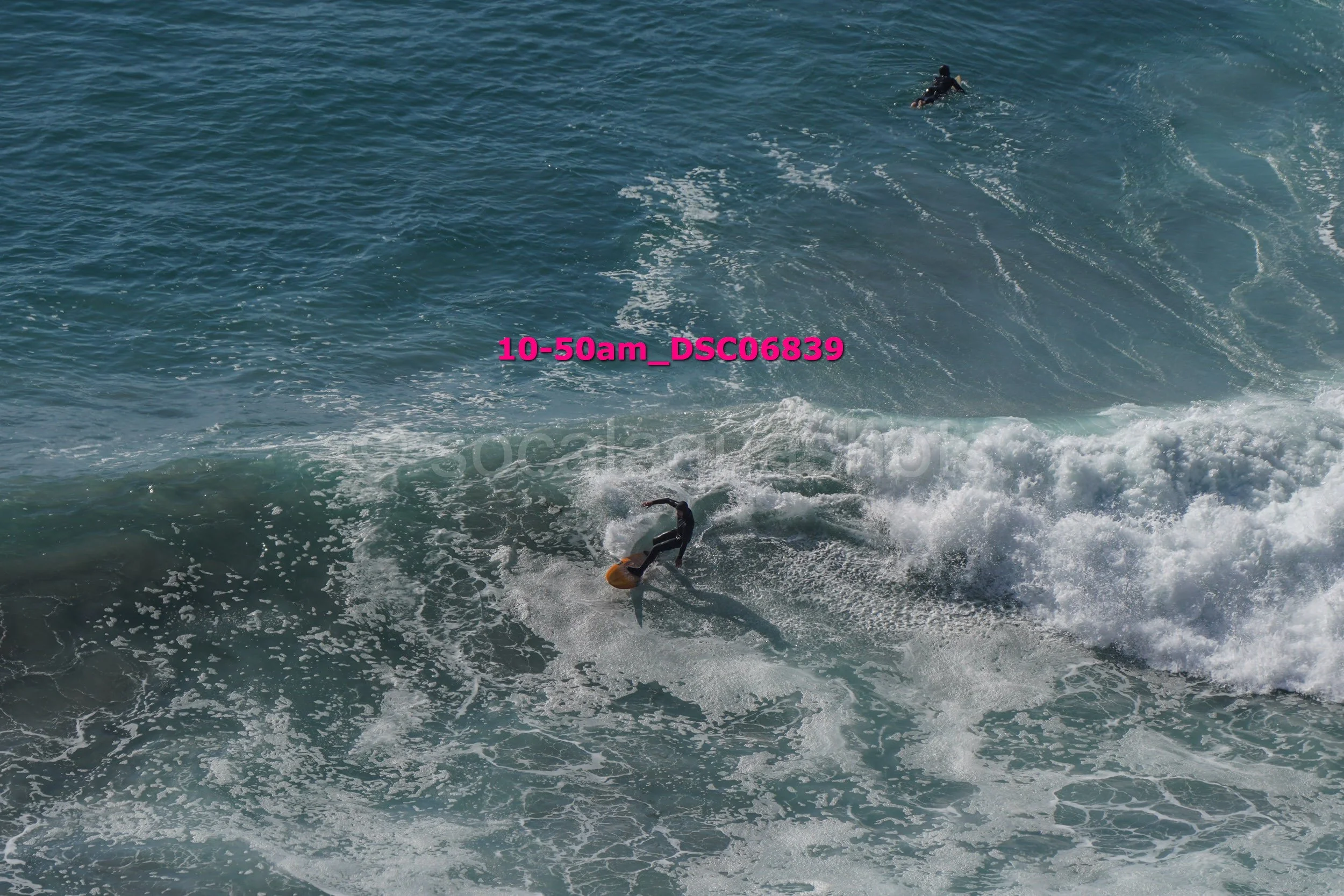 A person surfing on a wave in the ocean with a second surfer in the background. The water is blue with white foam from the breaking waves.