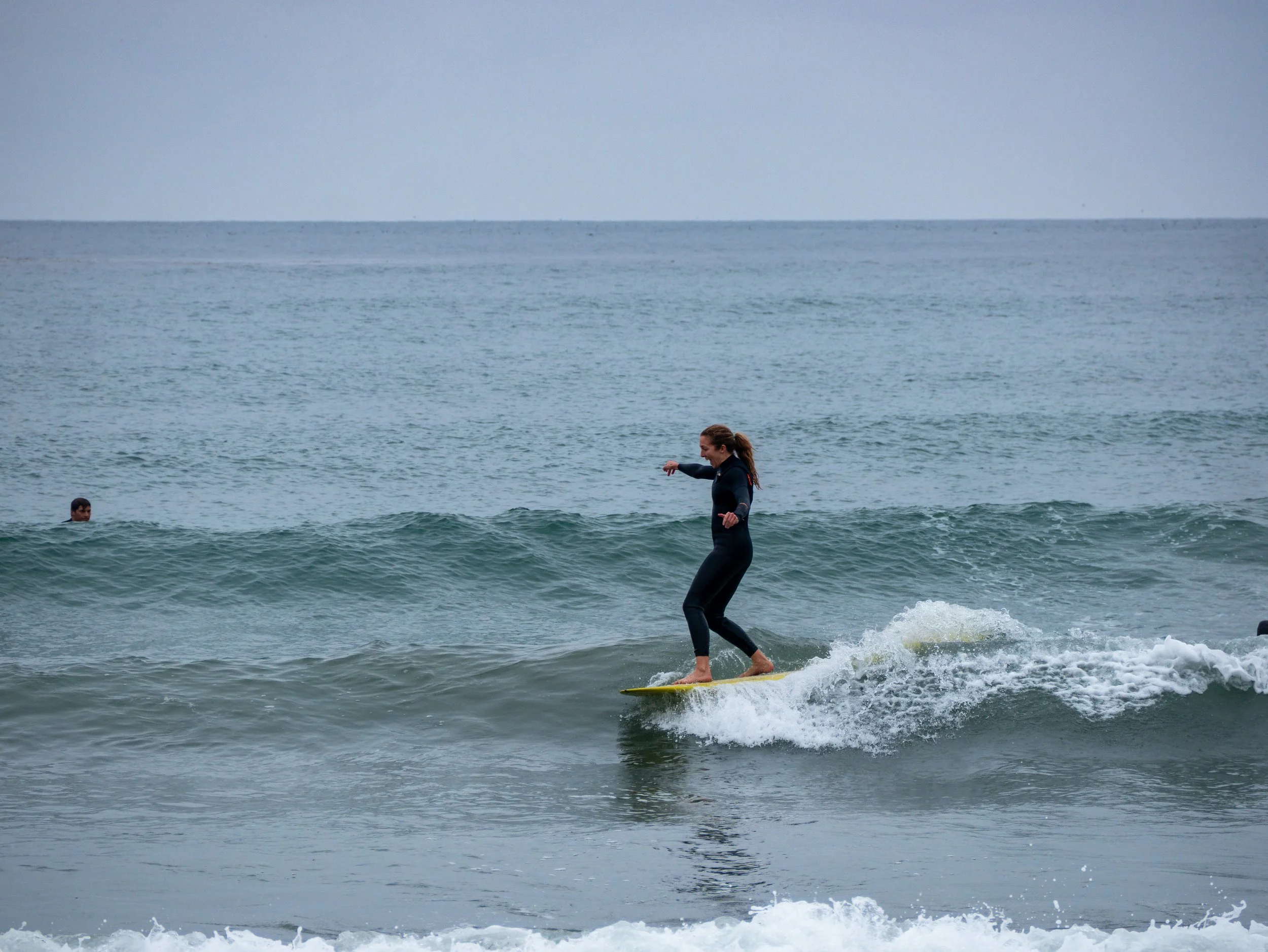 A woman surfing on a small wave in the ocean, wearing a black wetsuit, with a man swimming in the background.