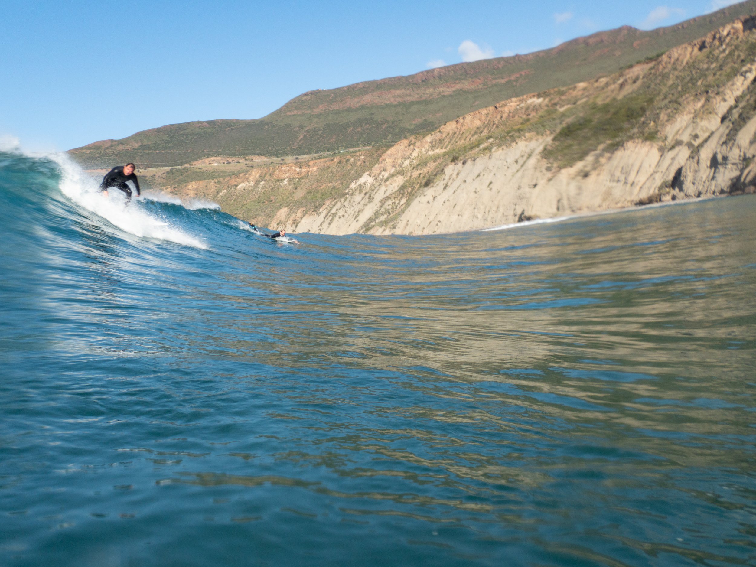 A person surfing on a blue wave near a rocky coastline with hills in the background.