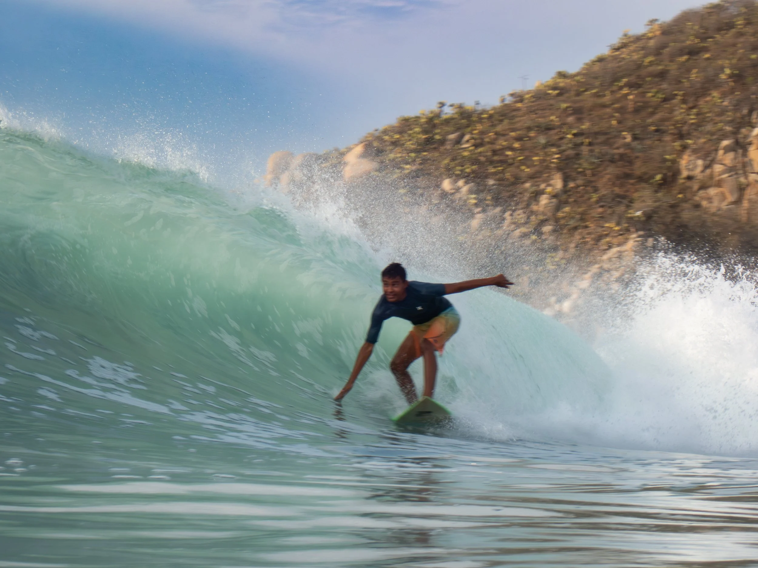 A person surfing on a turquoise wave near a rocky shoreline with sparse vegetation, during sunny weather.