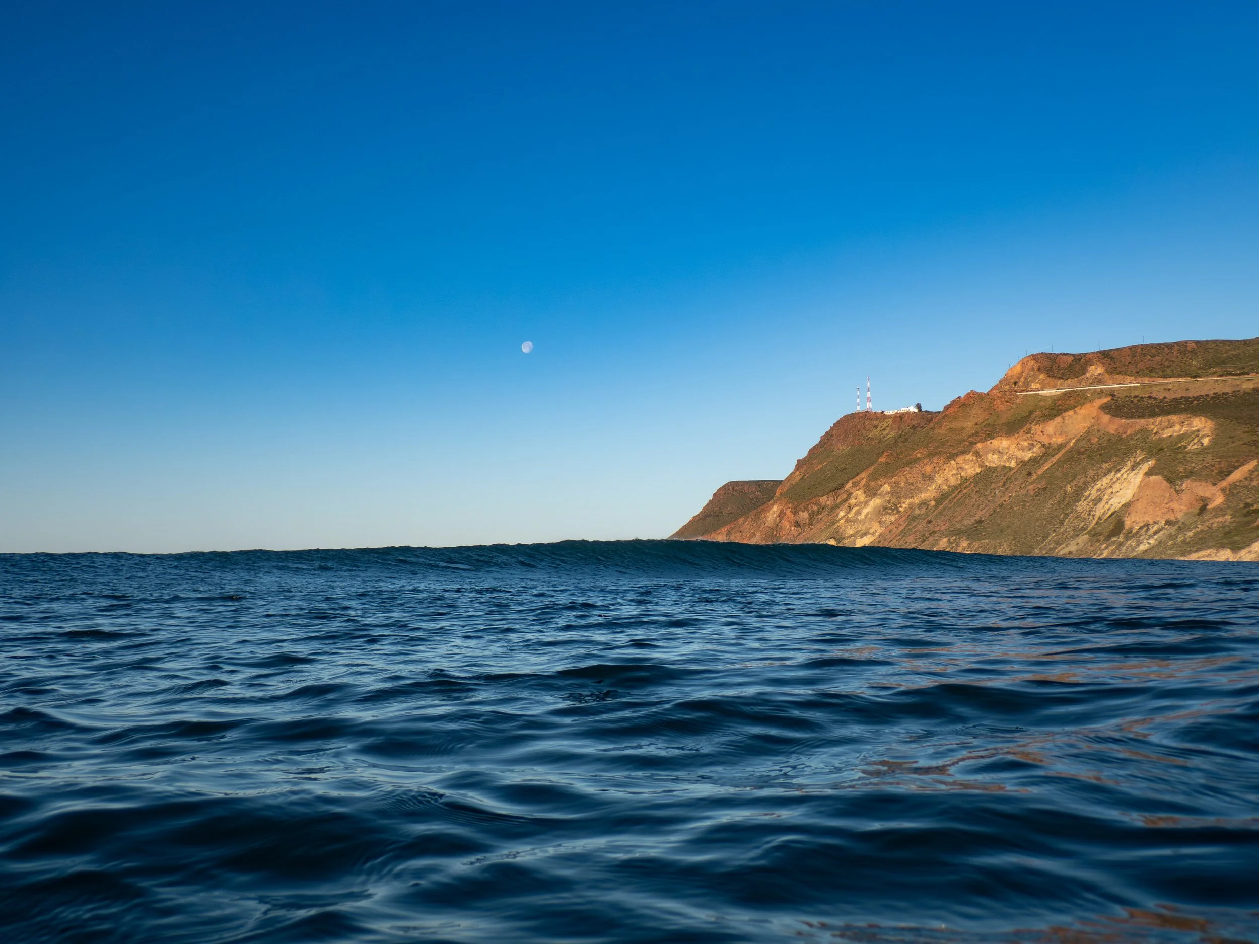 Ocean waves with a hillside in the background and a clear sky showing the moon.